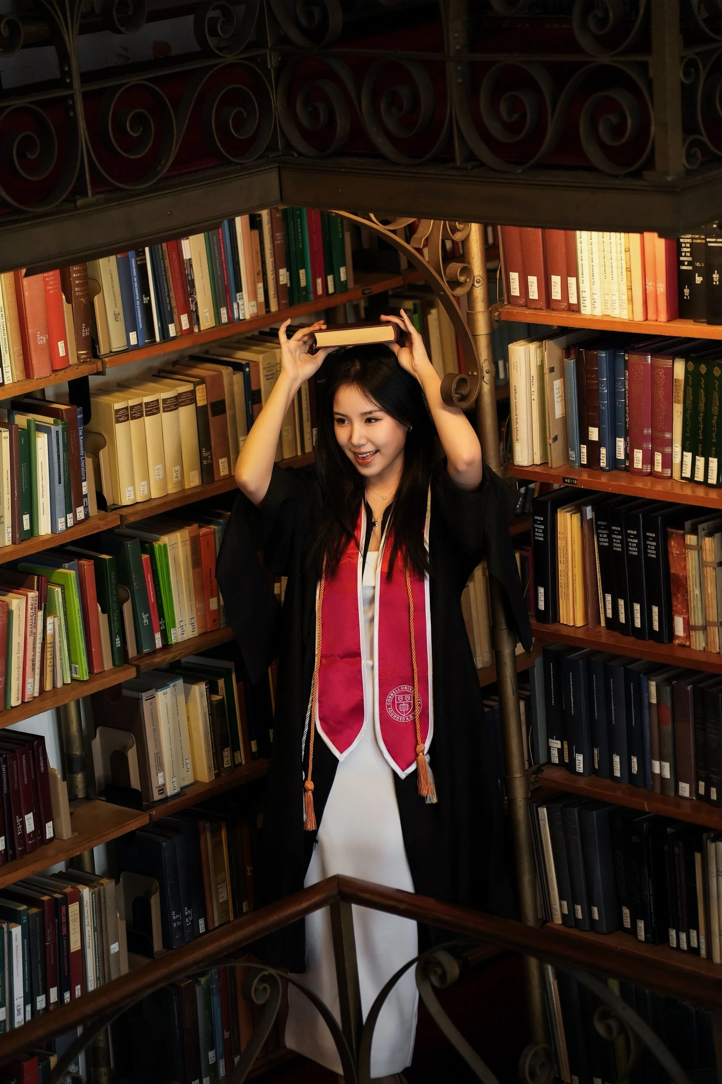 A young woman in a graduation gown and stole standing in a library, holding a book on her head, smiling, surrounded by bookshelves filled with books.