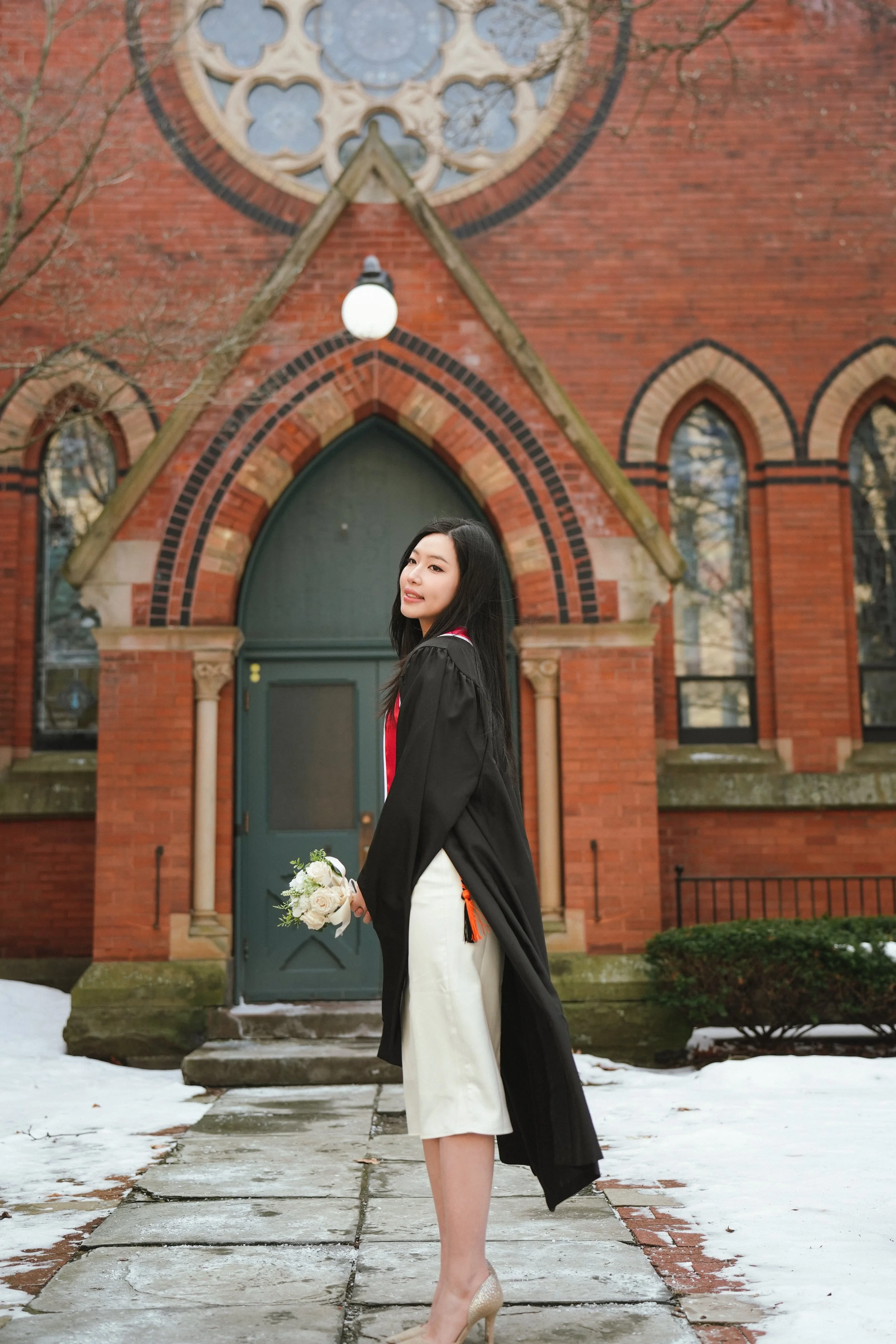 A young woman in a graduation gown holding a bouquet of white roses, standing on a stone pathway in front of a brick church with arched windows and a large round stained glass window, with some snow on the ground.