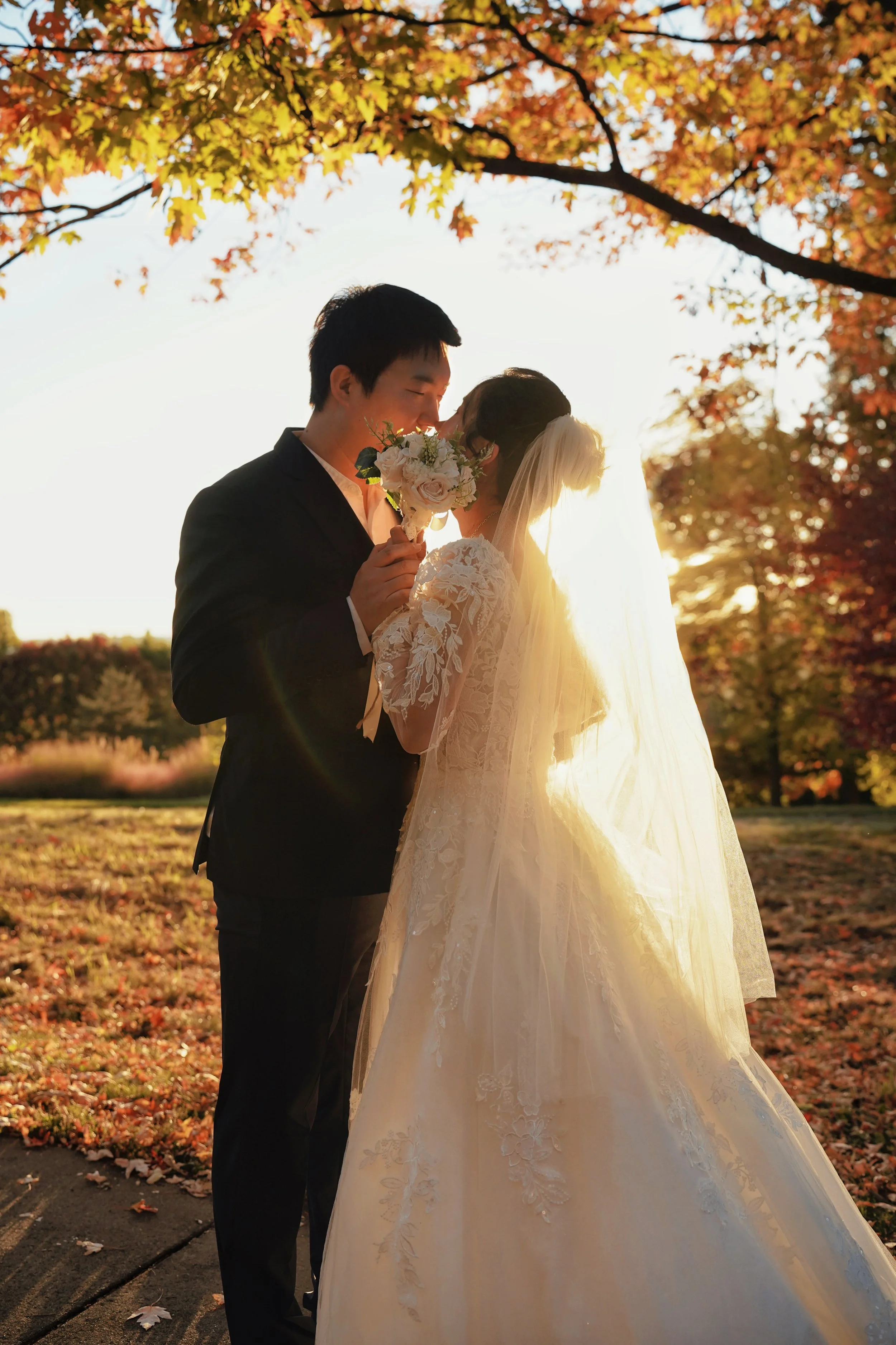 A bride and groom sharing a kiss outdoors during sunset, with autumn leaves on the trees in the background.