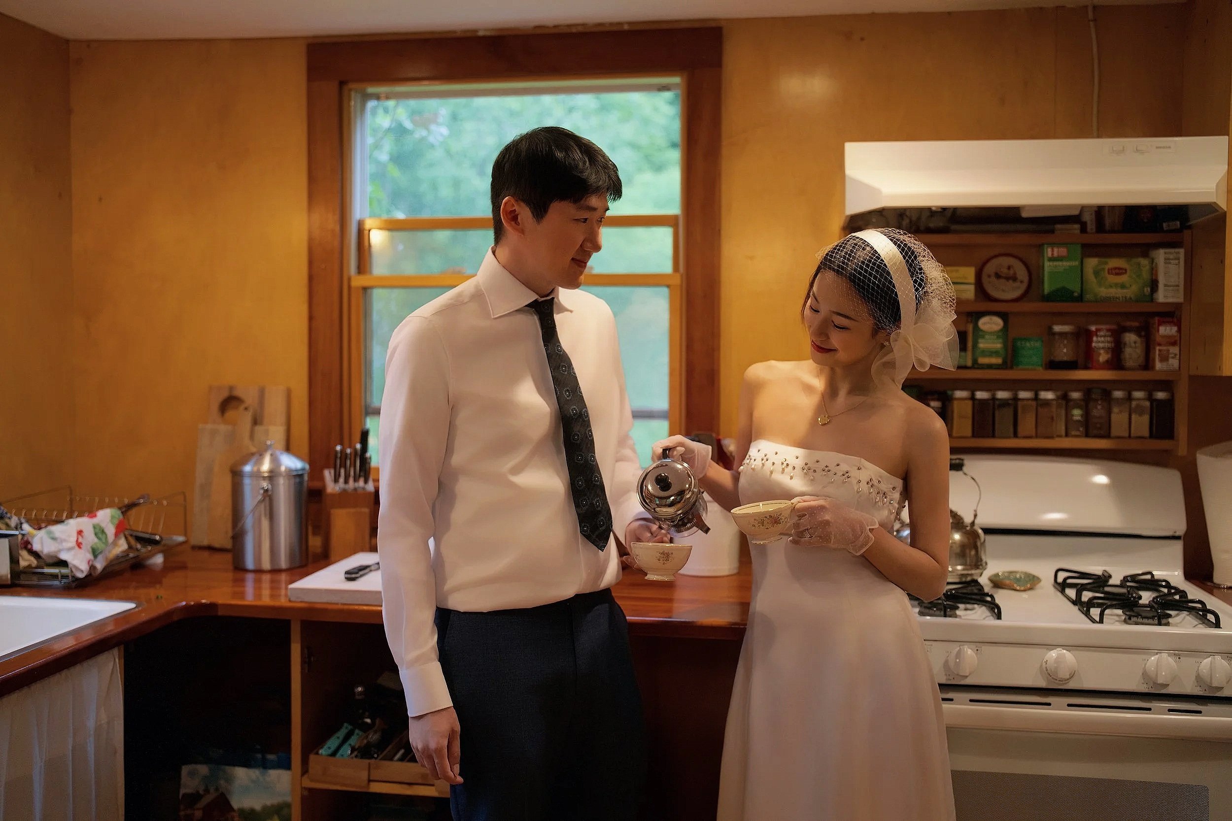 A couple in wedding attire sharing a moment in a cozy kitchen, with the woman holding a teacup and the man dressed in a tuxedo.