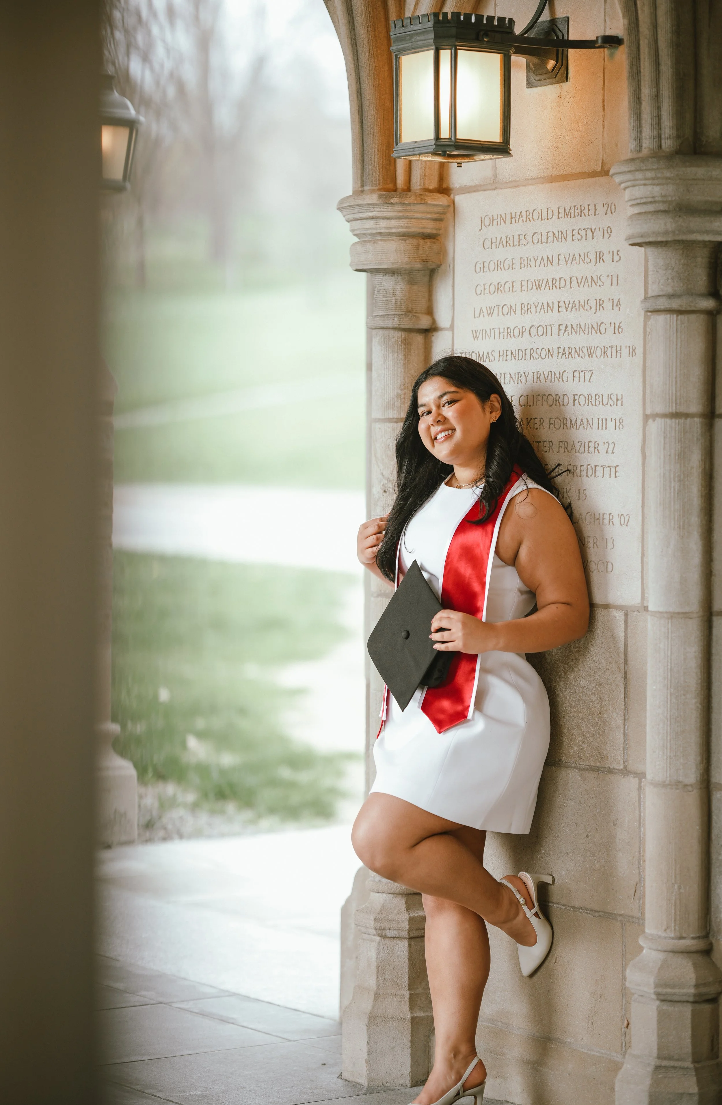 A young woman in a white dress and white heels is smiling and holding a graduation cap while leaning against a stone column at a graduation ceremony, with a grassy landscape and cloudy sky in the background.