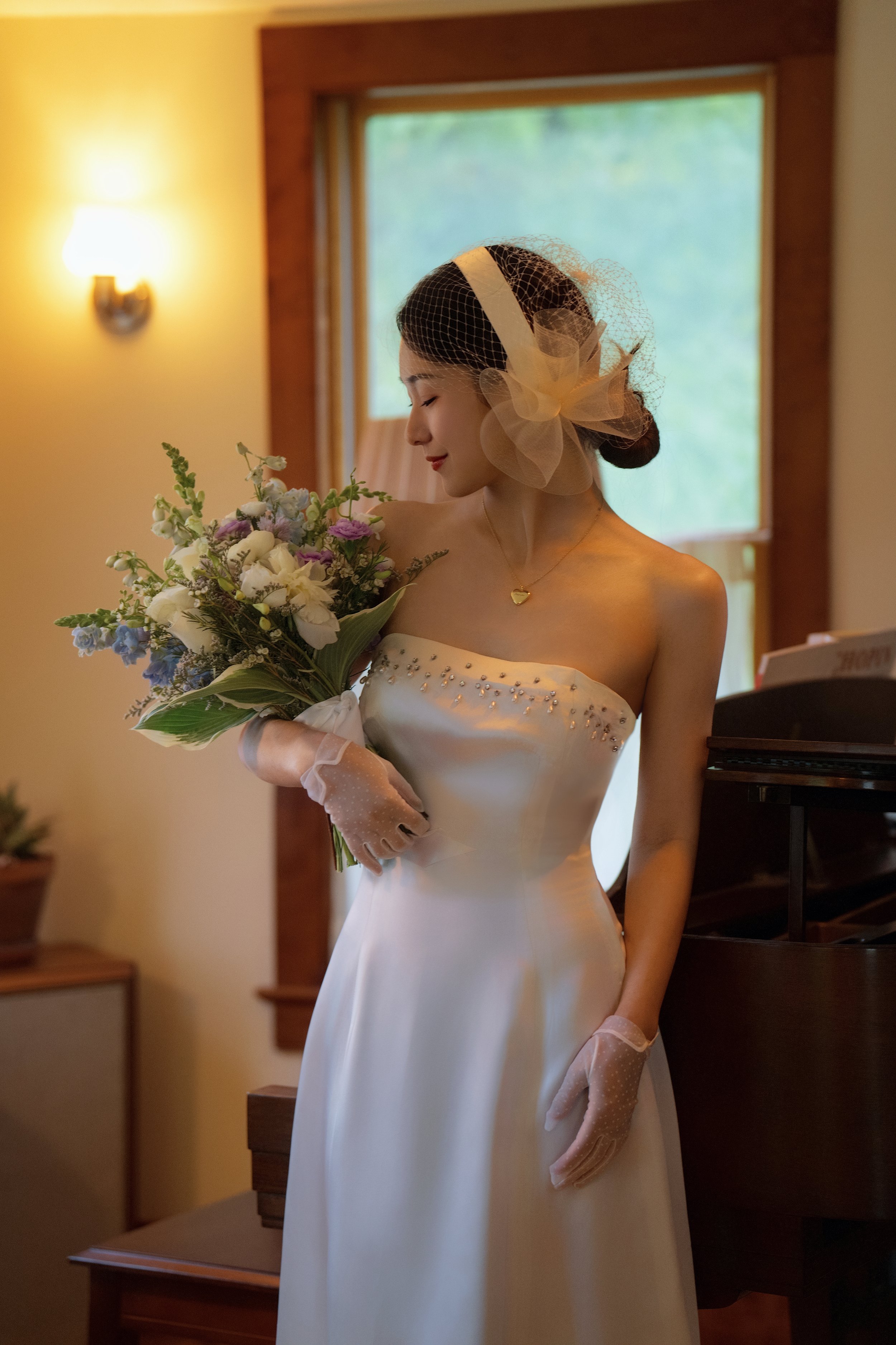 A bride in a strapless white wedding dress holds a bouquet of flowers, standing near a piano with soft lighting from a wall sconce and window.