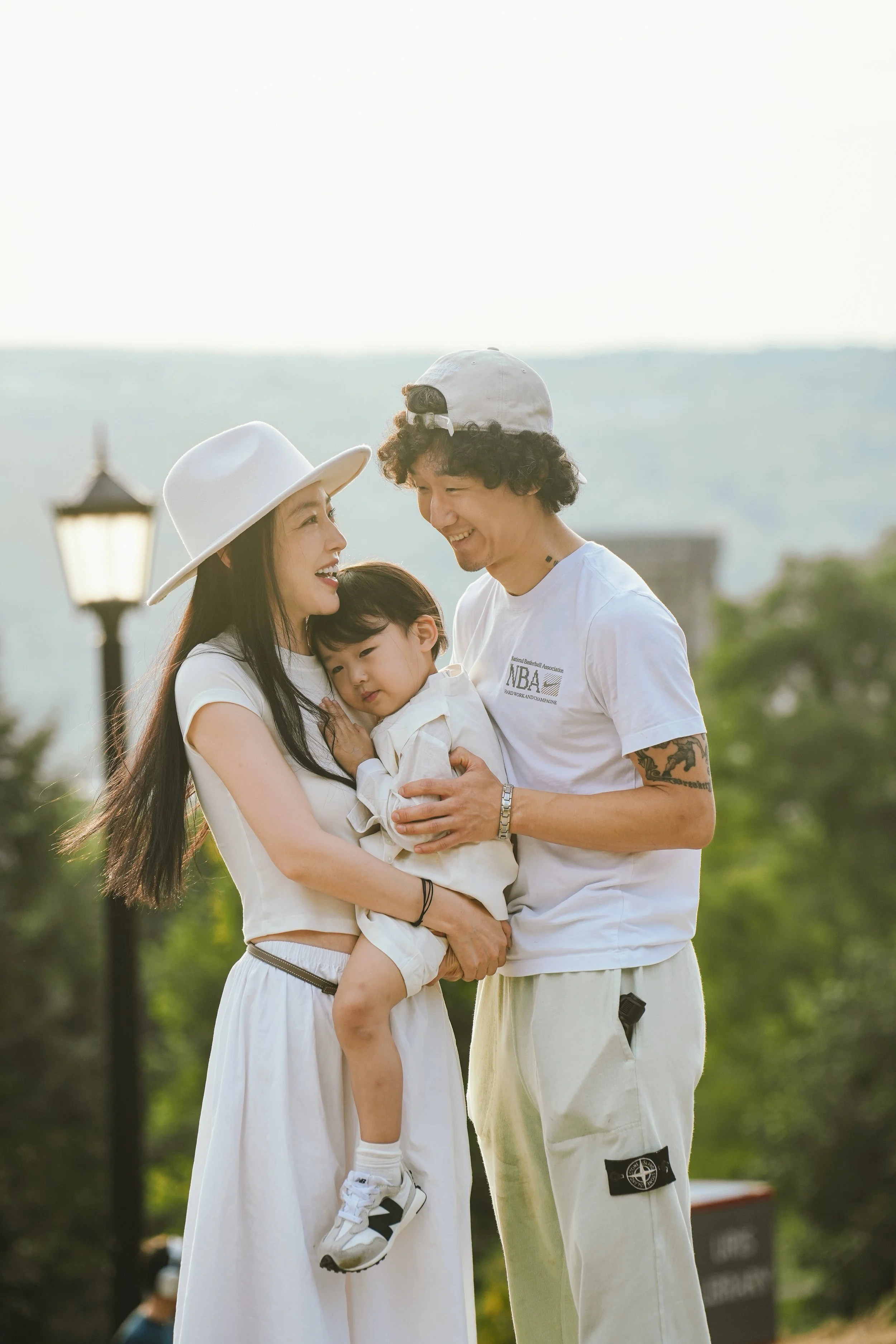 A family of three, an Asian woman, a Caucasian man, and a young child, standing outdoors and smiling at each other. The woman wears a white hat and skirt, the man a white cap and T-shirt, and the child is held in the woman's arms.