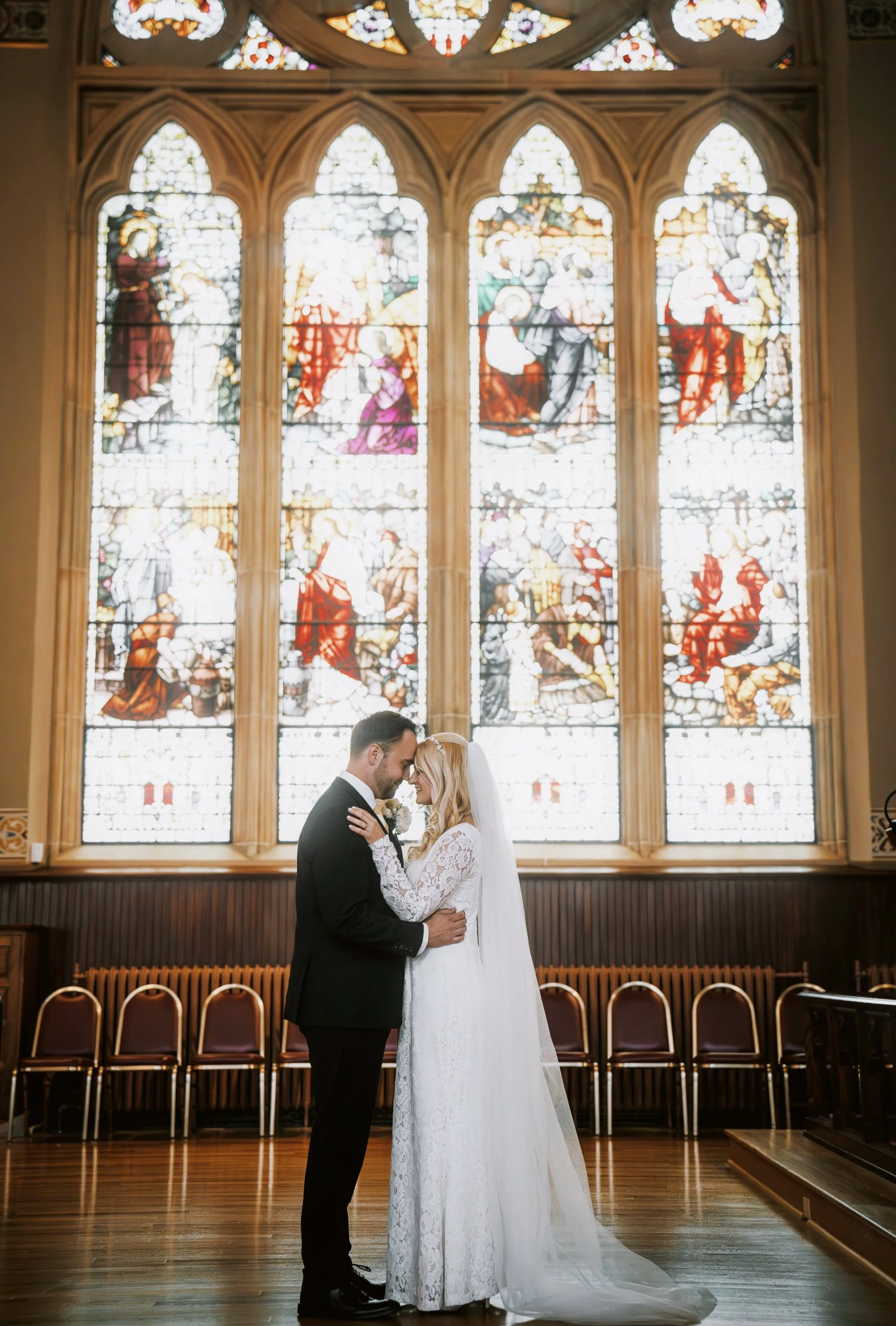 A bride and groom standing close together in front of a large stained glass window inside a church, embracing and touching foreheads.