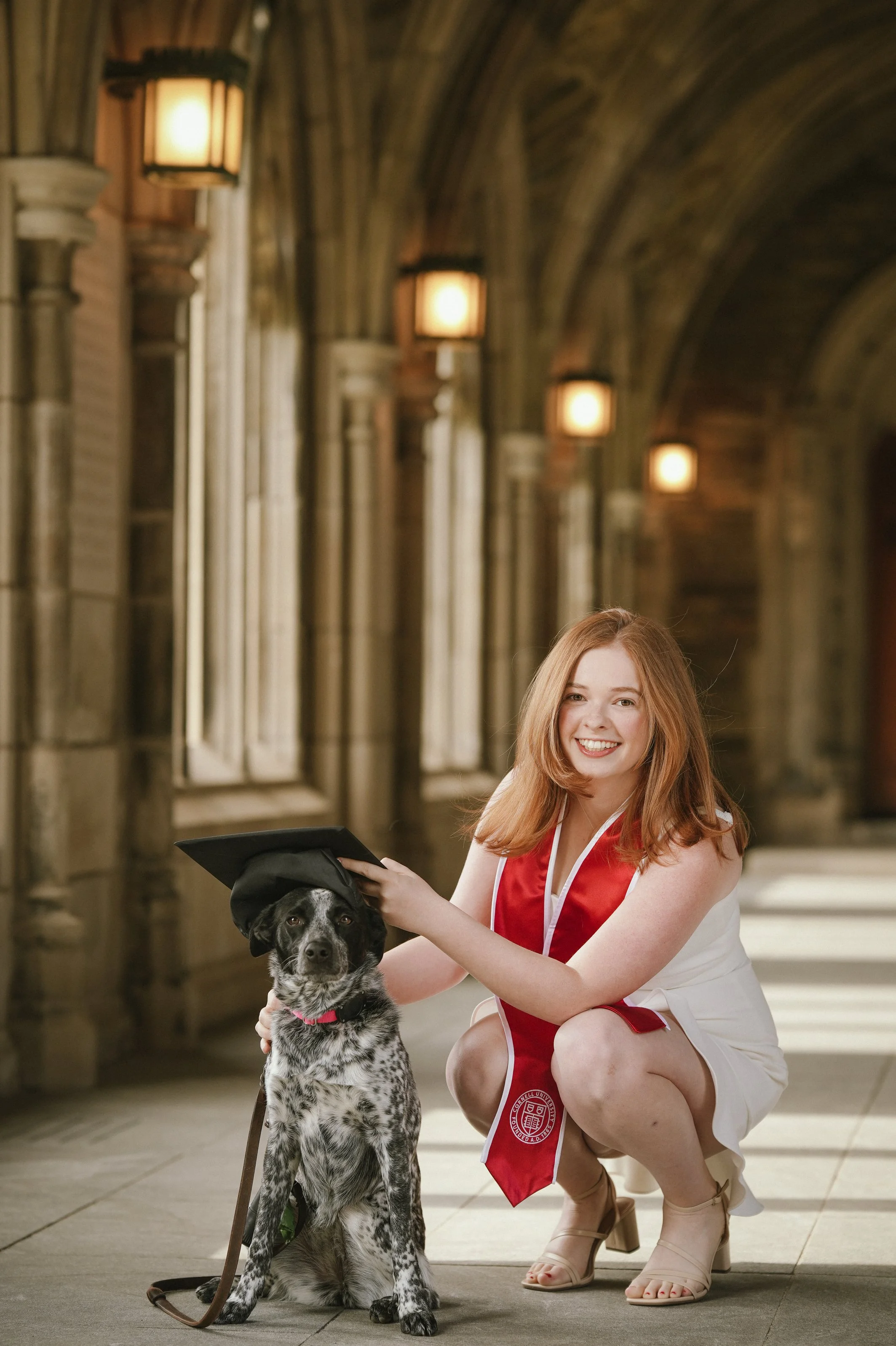 A young woman in a white dress and red graduation gown kneels next to a black and white dog holding a graduation cap, in an academic building with arched ceilings and wall-mounted lamps.