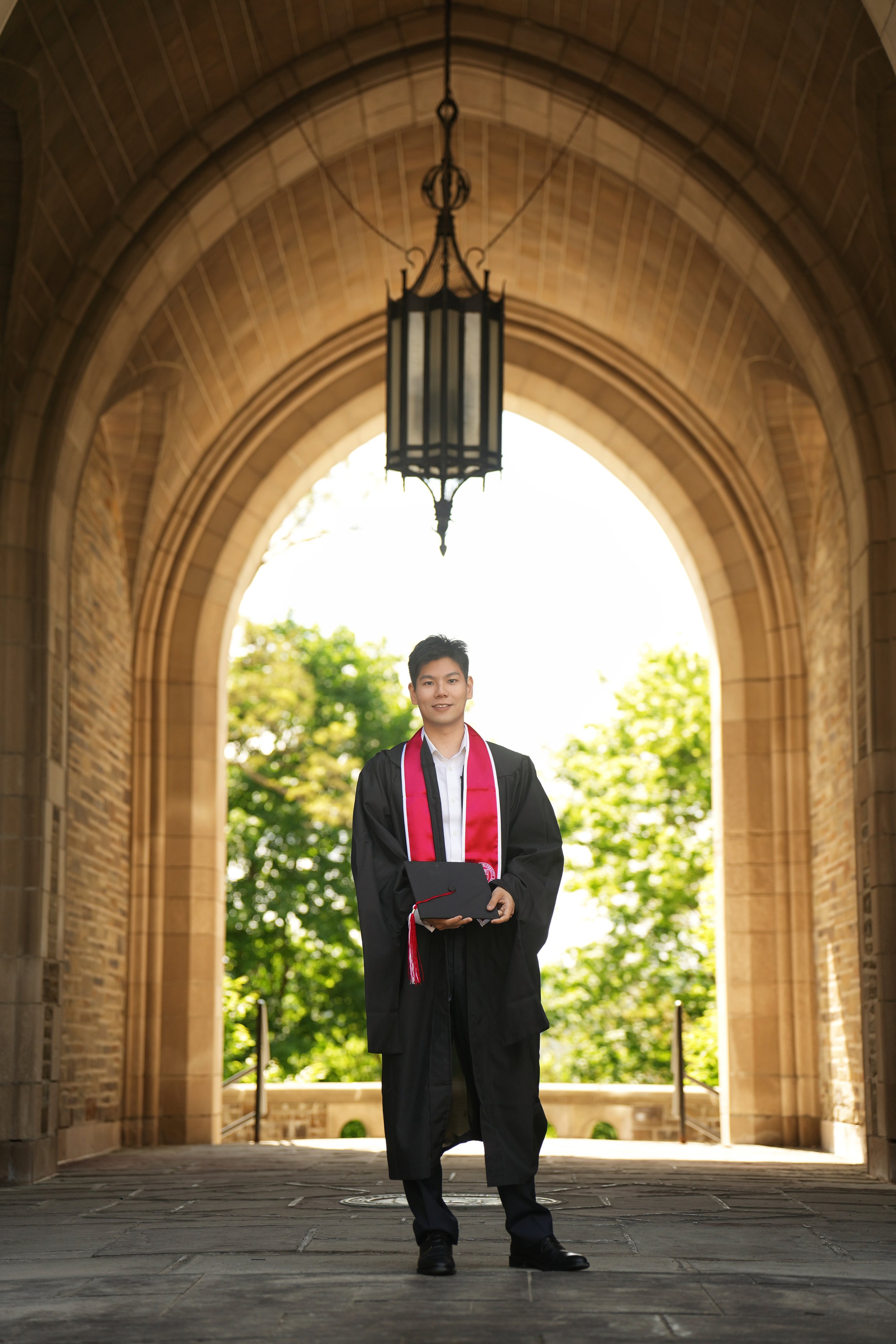 A young man in graduation gown and cap, wearing a red stole, holding a diploma, standing under an arched stone structure with a hanging lantern, outdoors with green trees in the background.