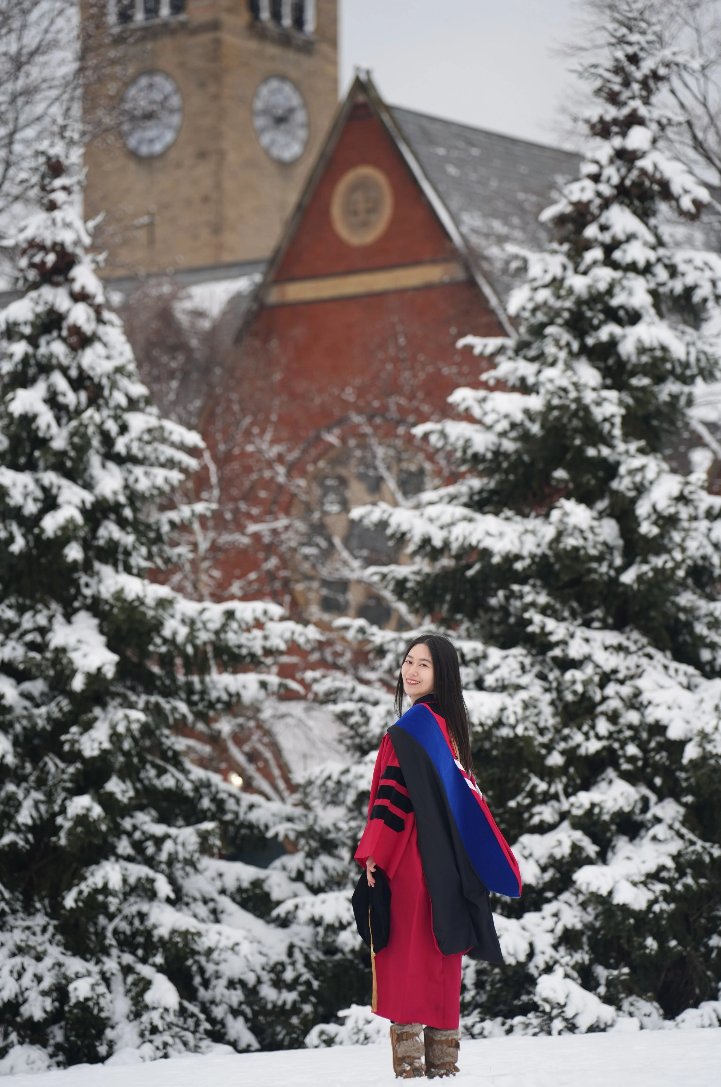 Young woman in graduation gown and stole, holding a cap, standing on snow with snow-covered trees and a church in the background.
