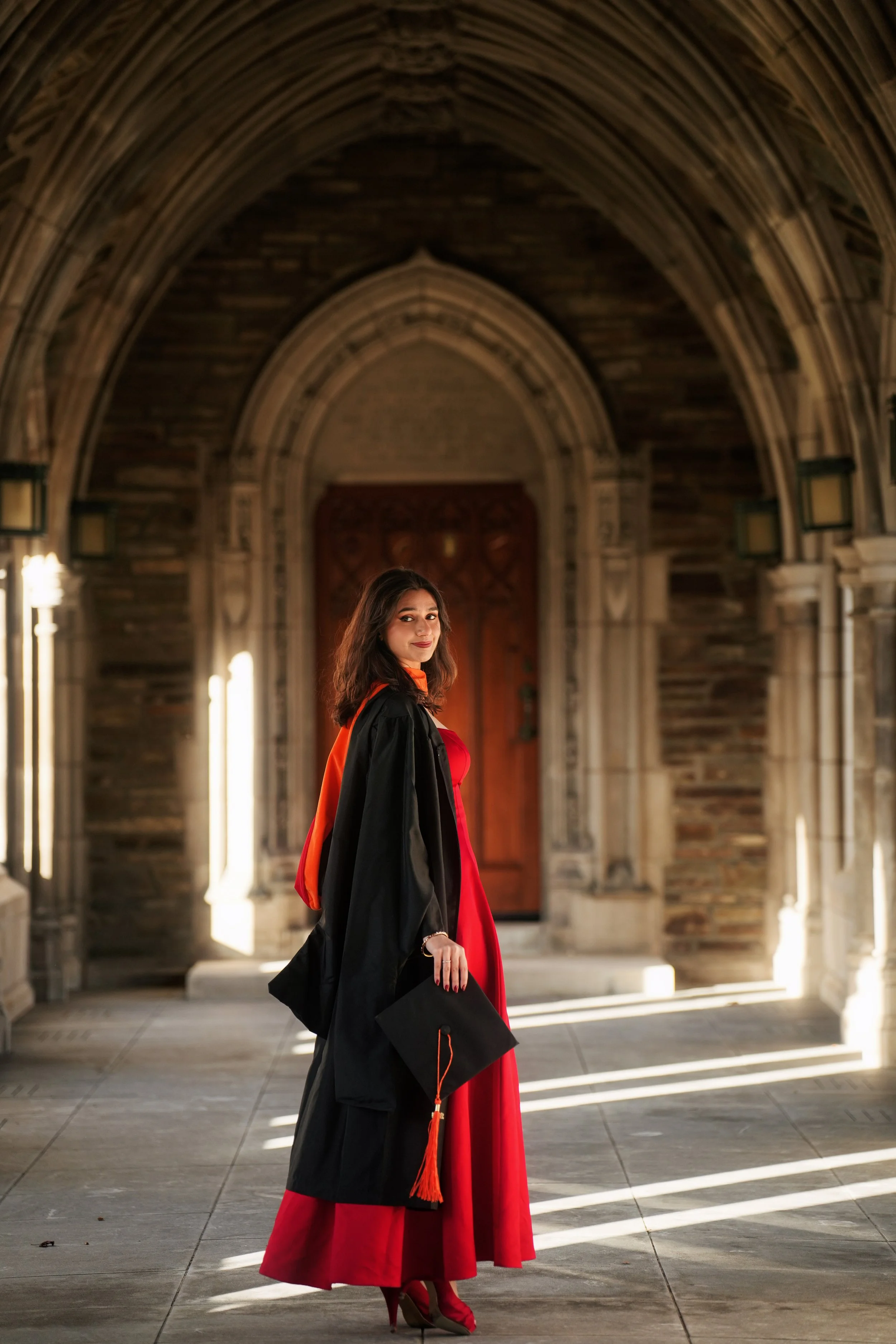 A young woman in a red dress and high heels holding a graduation cap stands in a stone archway with sunlight streaming through.