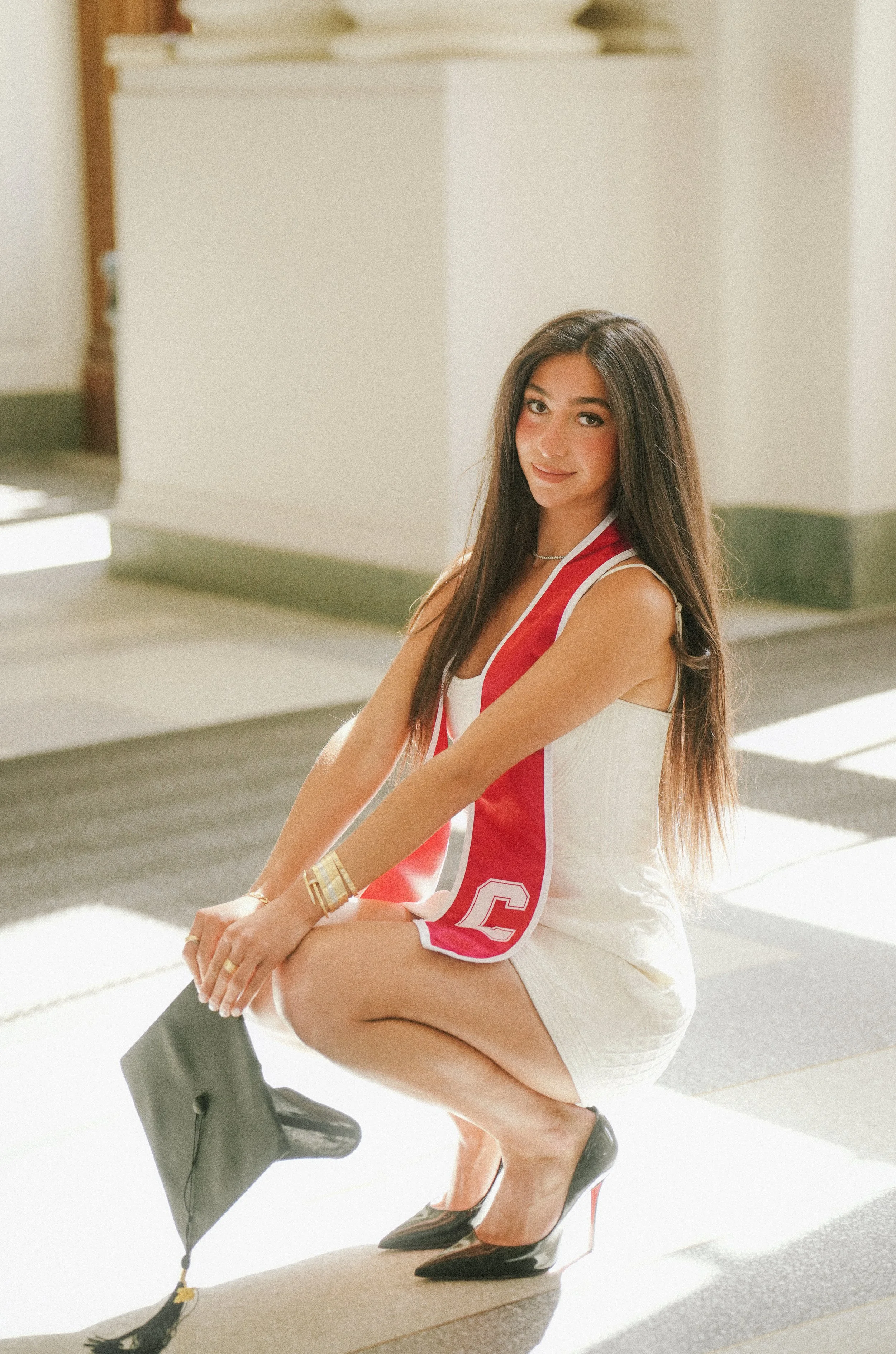 A young woman with long dark hair, wearing a white sleeveless dress with red accents, black high heels, and gold jewelry, sitting on a step in a sunlit indoor space with a white wall background.