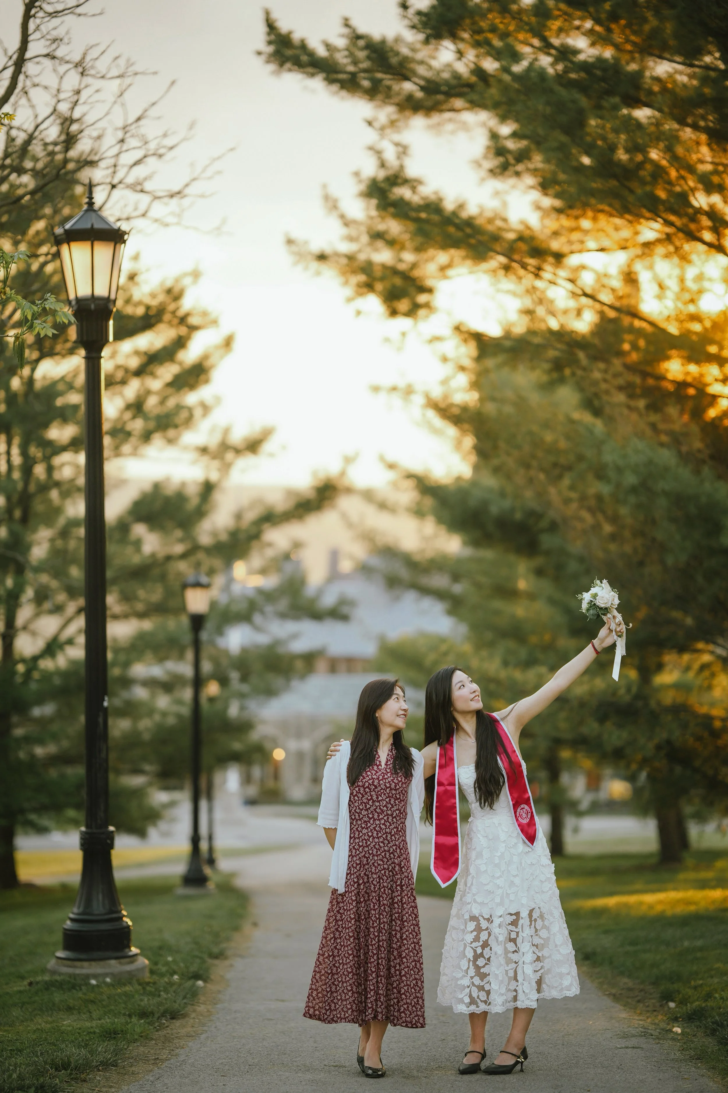 Two women are walking along a park path during sunset, one in a white lace dress with a sash and the other in a red patterned dress with a white cardigan. The woman in the white dress is holding a bouquet of flowers high in the air, and they are smil