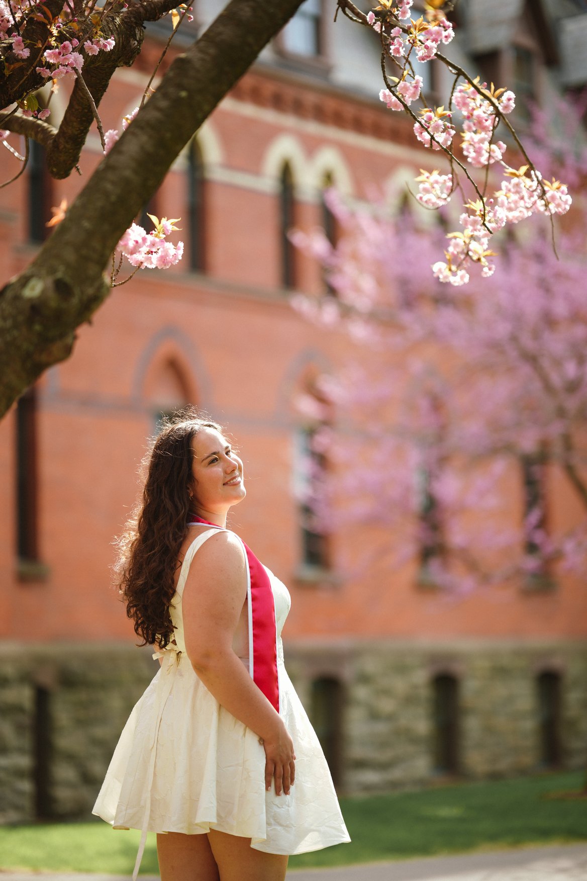 A woman with long curly brown hair, wearing a sleeveless cream-colored dress with a red sash, stands outdoors with a smile and looks up. She is surrounded by pink blooming cherry blossom trees, and a red brick building with arched windows is in the b