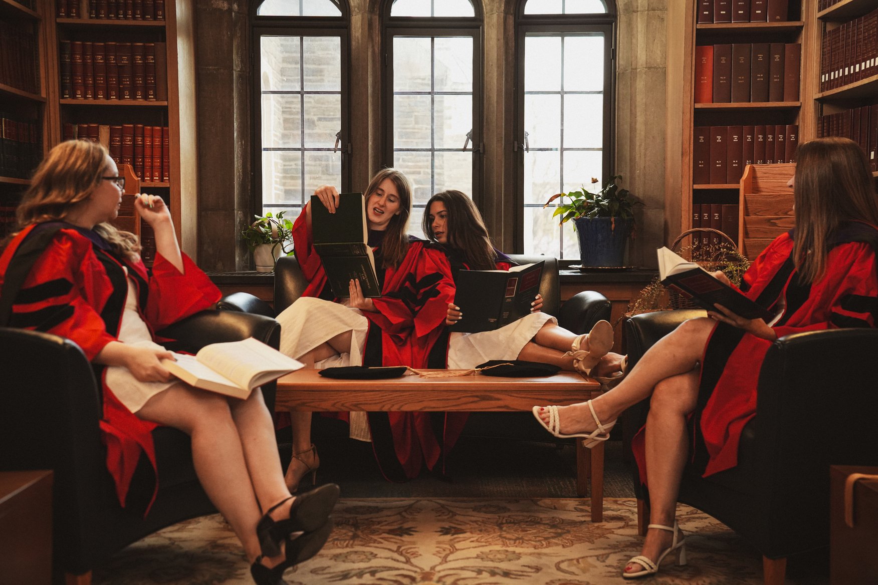 Group of five women in red and black graduation gowns sitting and relaxing in a cozy library lounge with windowseat, reading books and engaging in conversation.