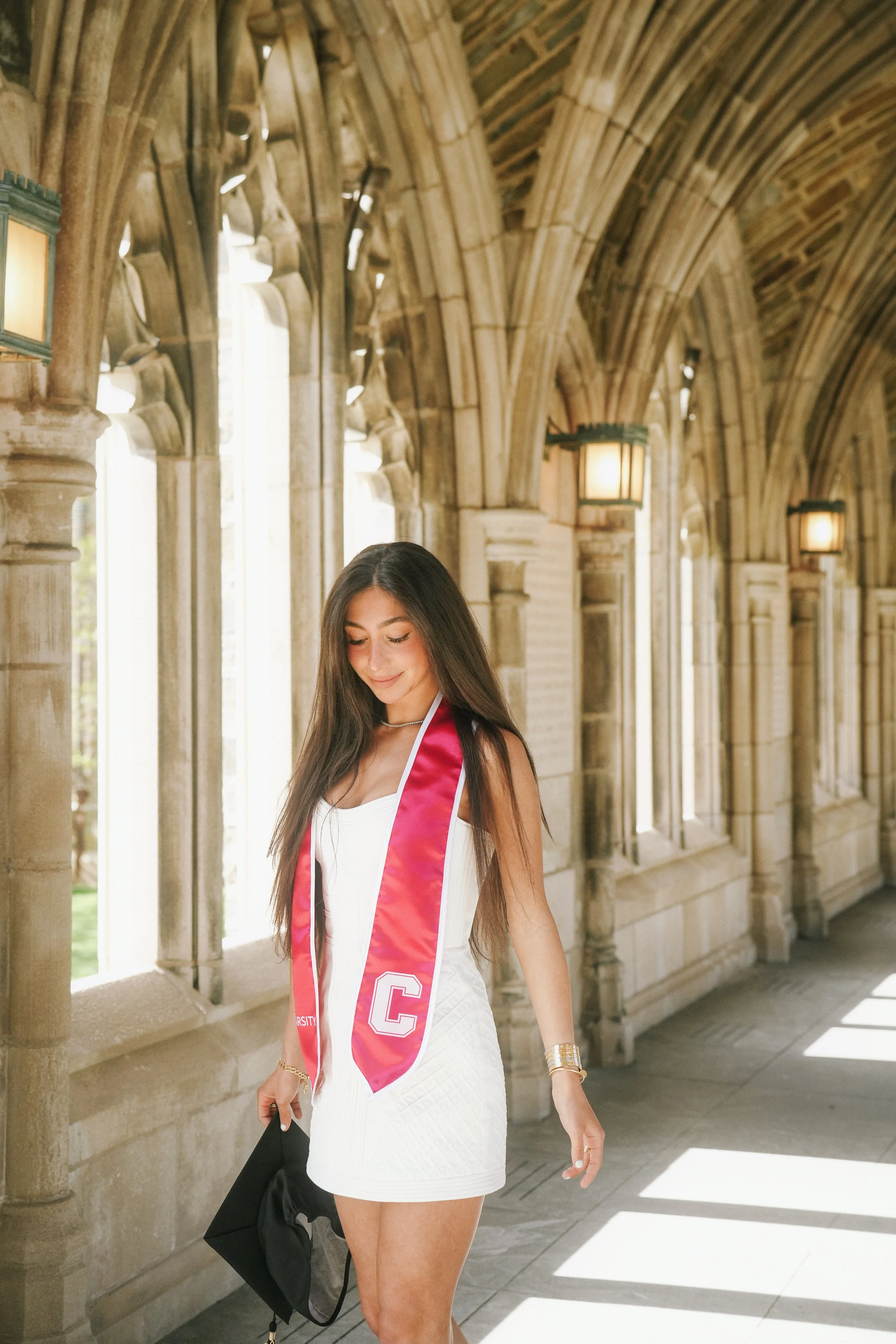 Young woman in a white dress with a red graduation stole, holding a black bag, walking in a stone arch corridor with hanging lights.