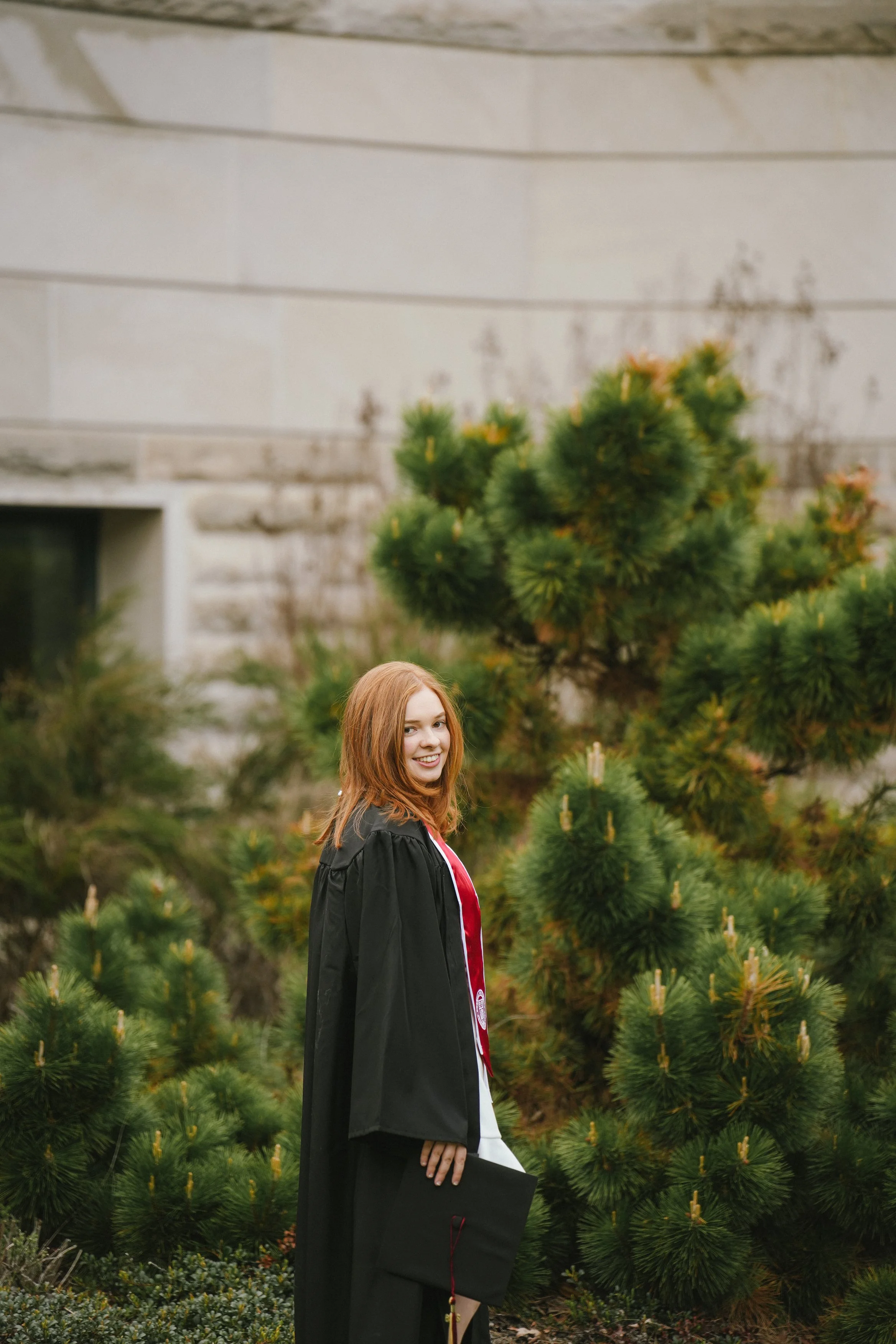 A young woman with red hair smiling in graduation gown holding a diploma in front of a green pine tree outdoors.