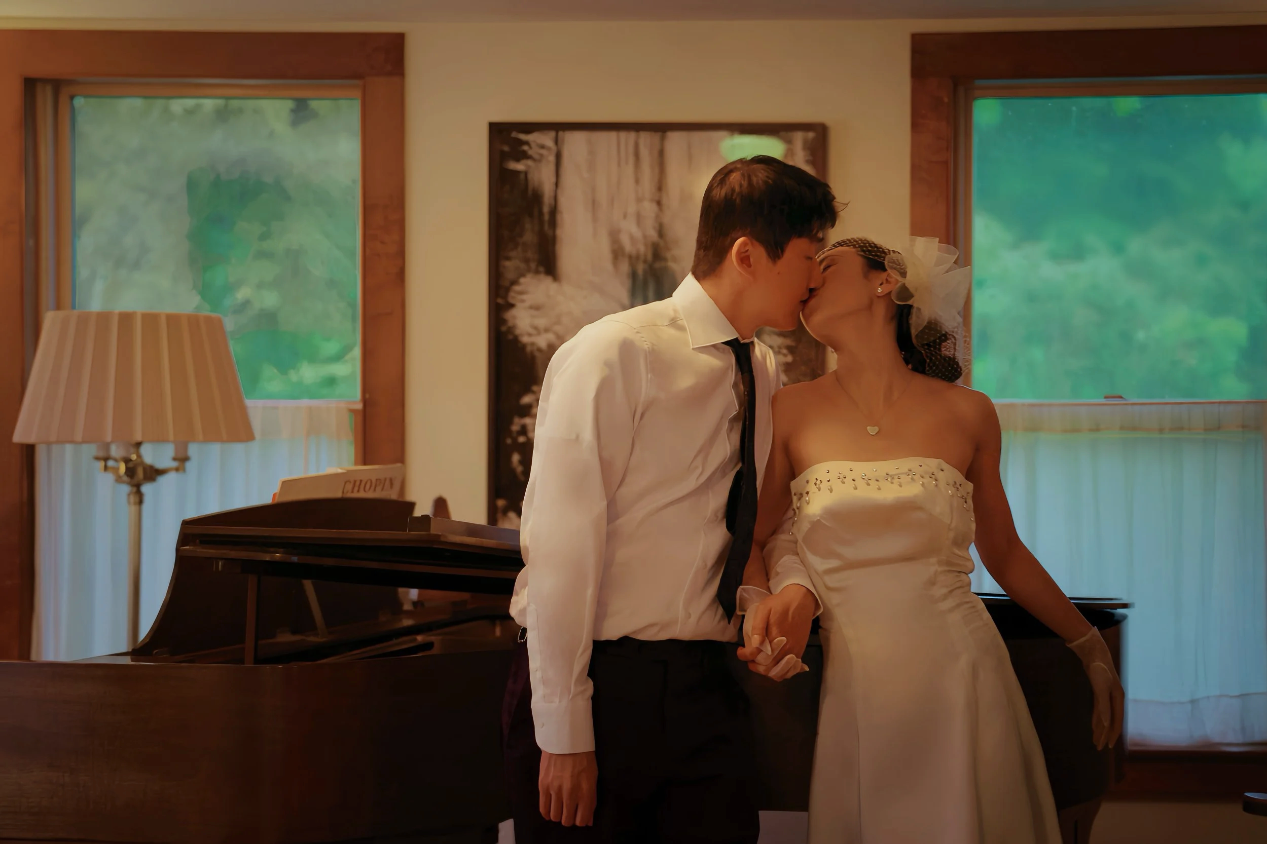 A couple sharing a kiss in a cozy, warmly lit room with large windows showing green trees outside.
