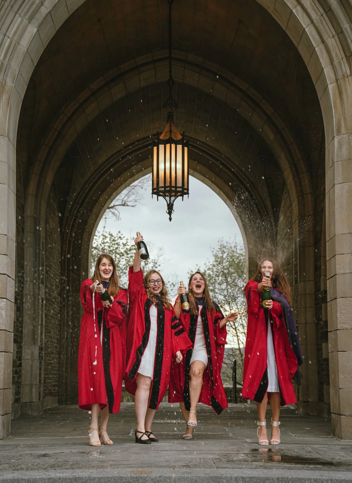 Four women celebrating graduation under an archway, wearing red graduation gowns, holding champagne bottles, and smiling.