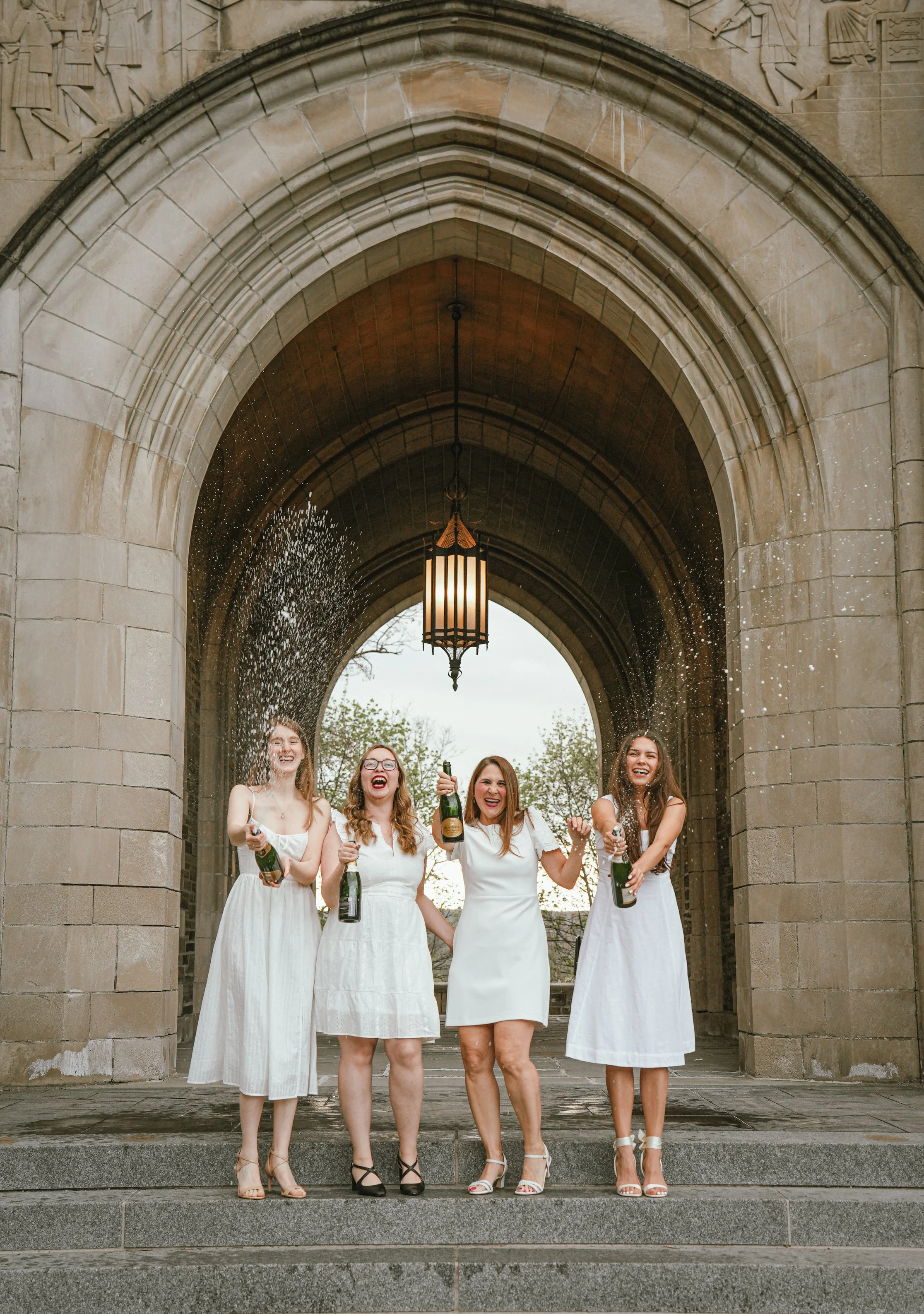 Five women in white dresses celebrating with champagne under a stone archway, smiling and spraying champagne.