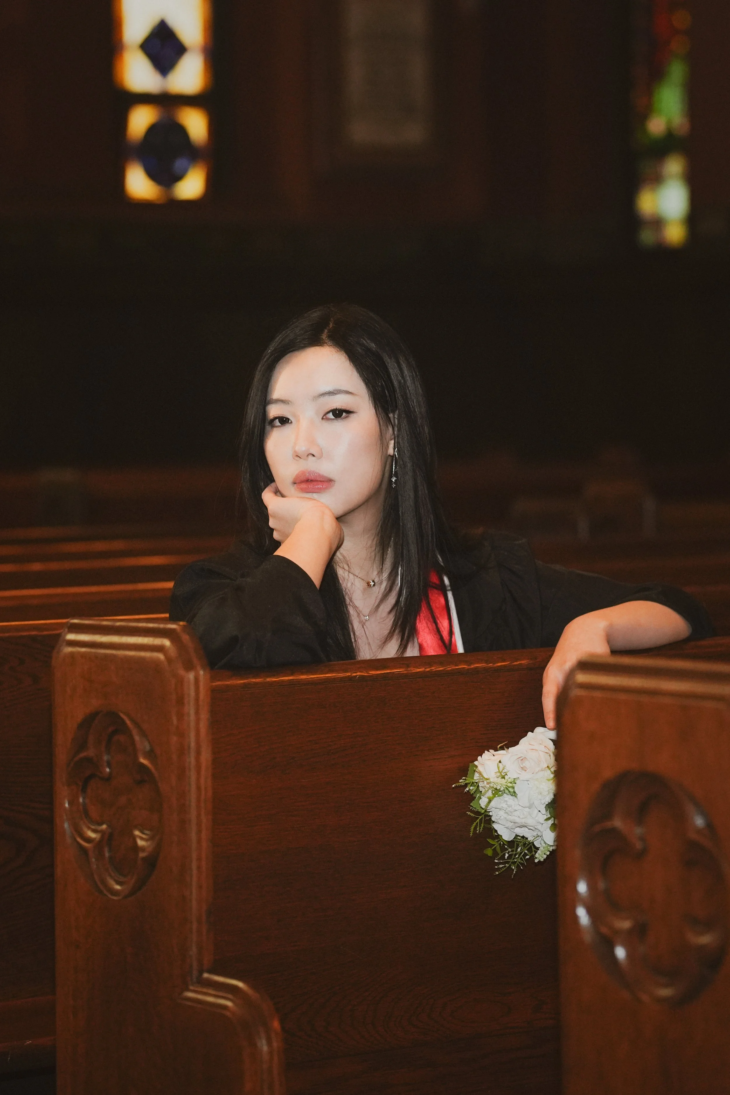 A woman with long black hair sitting in a church pew, resting her chin on her hand, with a bouquet of white flowers in front of her, inside a church with stained glass windows.
