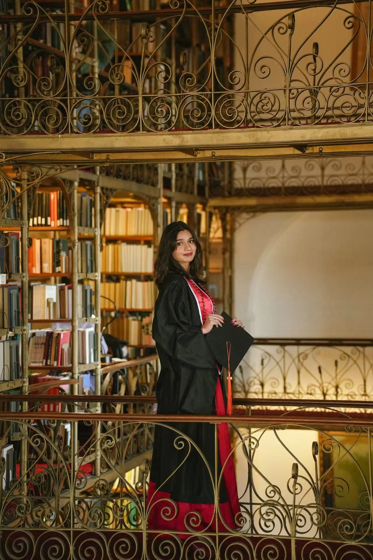A young woman in a graduation gown and cap holding a diploma, standing on a balcony in a library filled with books.