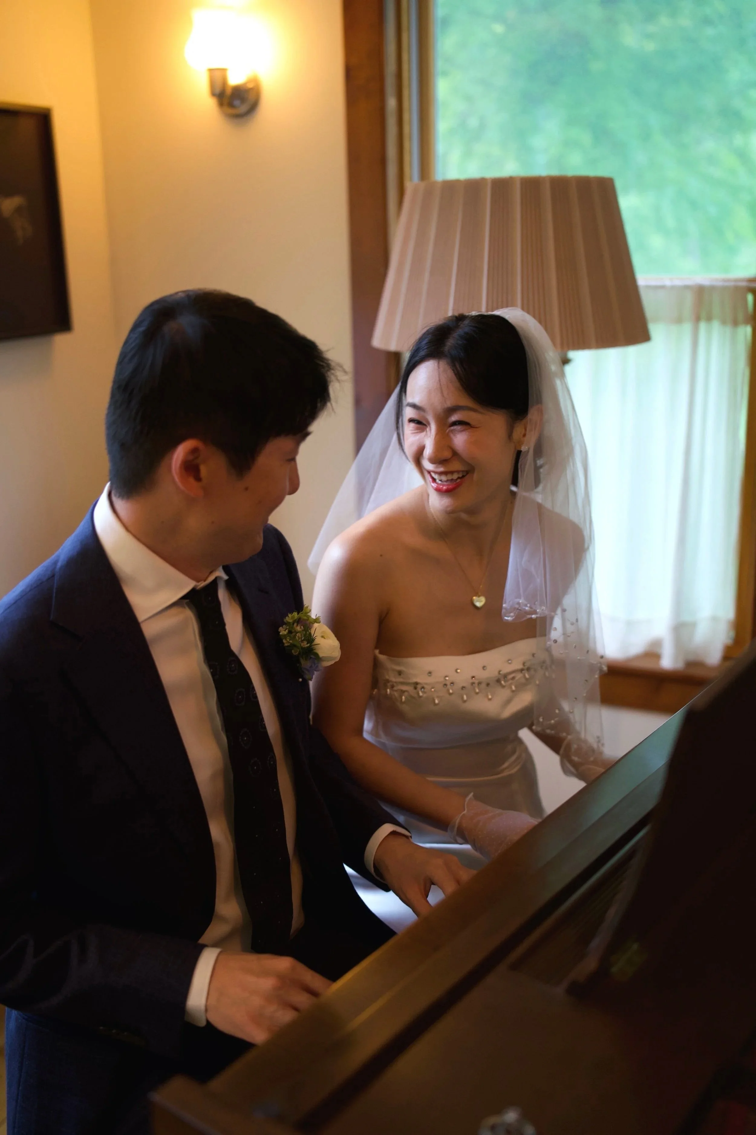 A bride in a white wedding dress and veil is smiling and singing along while playing the piano with a groom in a dark suit and tie, surrounded by warm indoor lighting and a window with greenery outside.