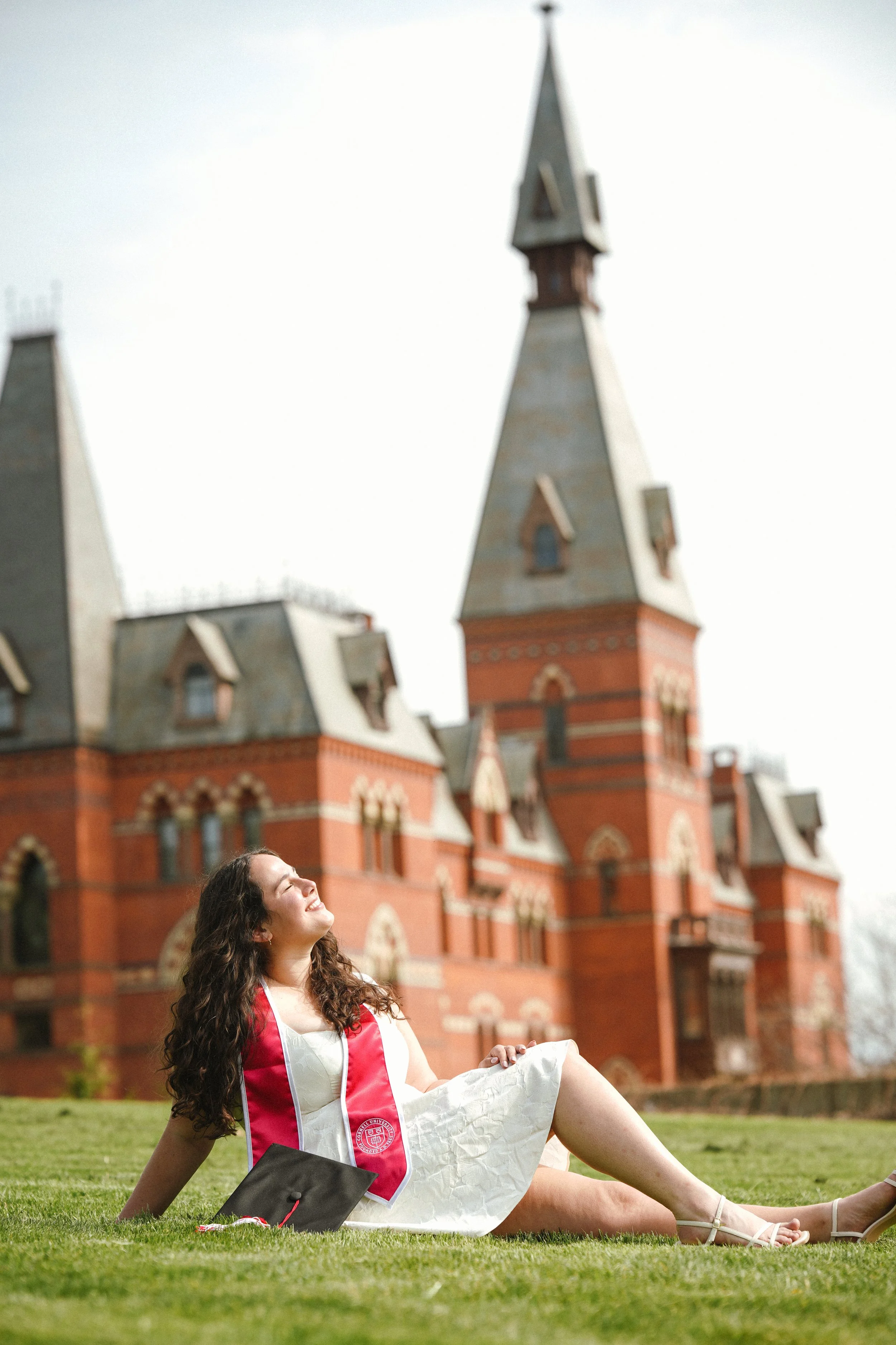 A young woman with long curly hair, wearing a white dress and a red graduation stole, sitting on grass, smiling and looking up, with a large brick and stone Gothic-style building in the background.
