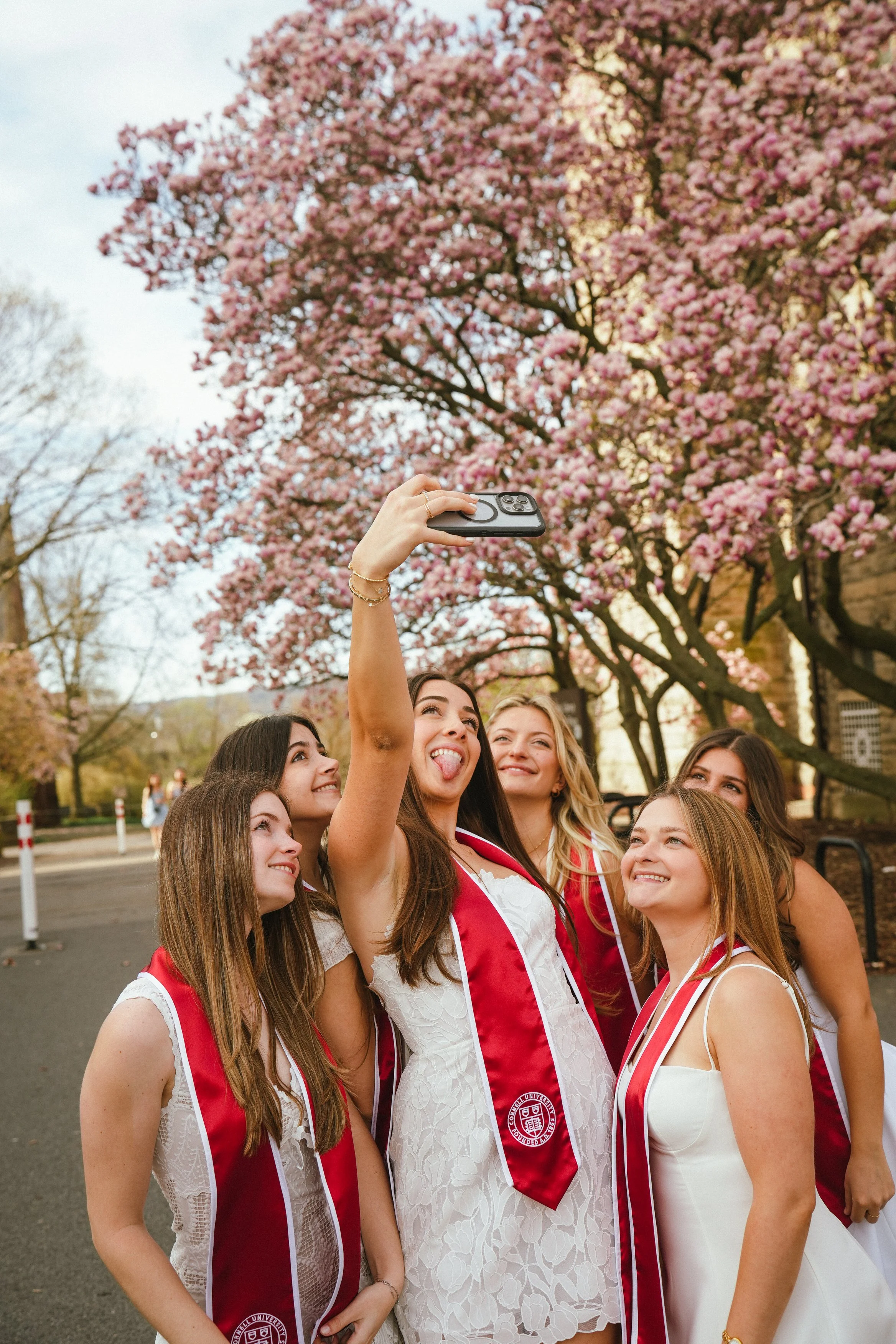 A group of young women taking a selfie in front of pink blossoming trees, wearing white dresses with red and white stoles that have a university emblem, outdoors during spring.