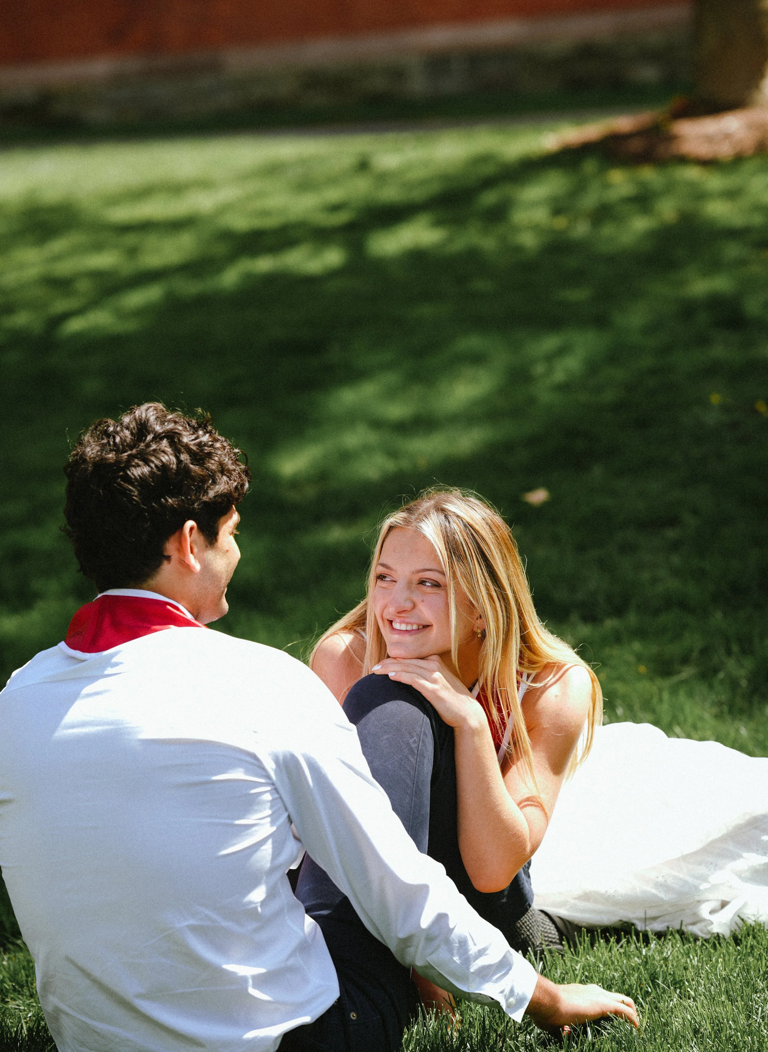 A young man and woman sitting on the grass outdoors, engaging in conversation and smiling at each other. The man has dark curly hair and is dressed in a white shirt with a red collar, while the woman has long blonde hair and is wearing a red tank top