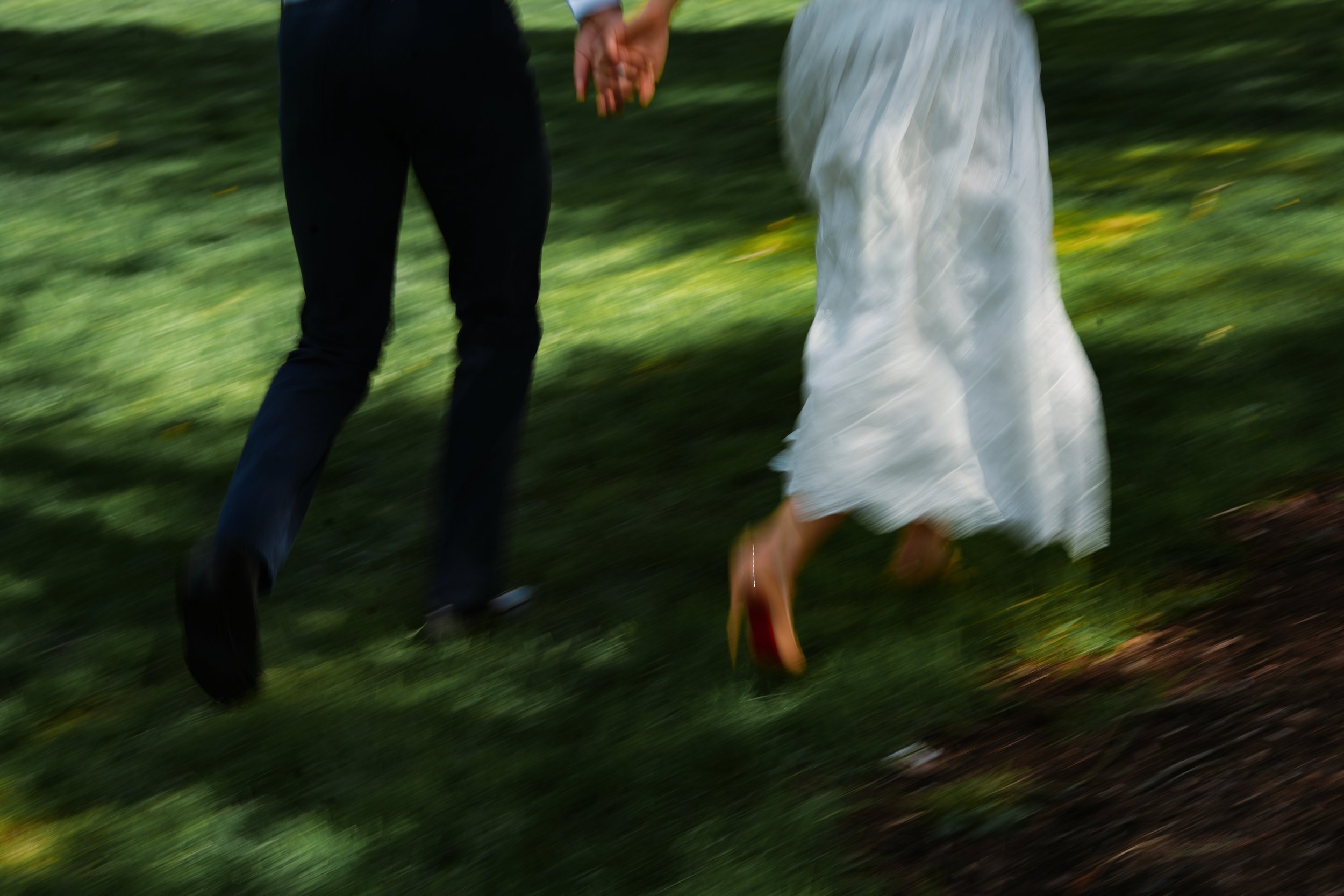 Close-up of a couple holding hands while walking outdoors on grass, with the woman wearing a white dress and shoes and the man dressed in dark pants and shirt.