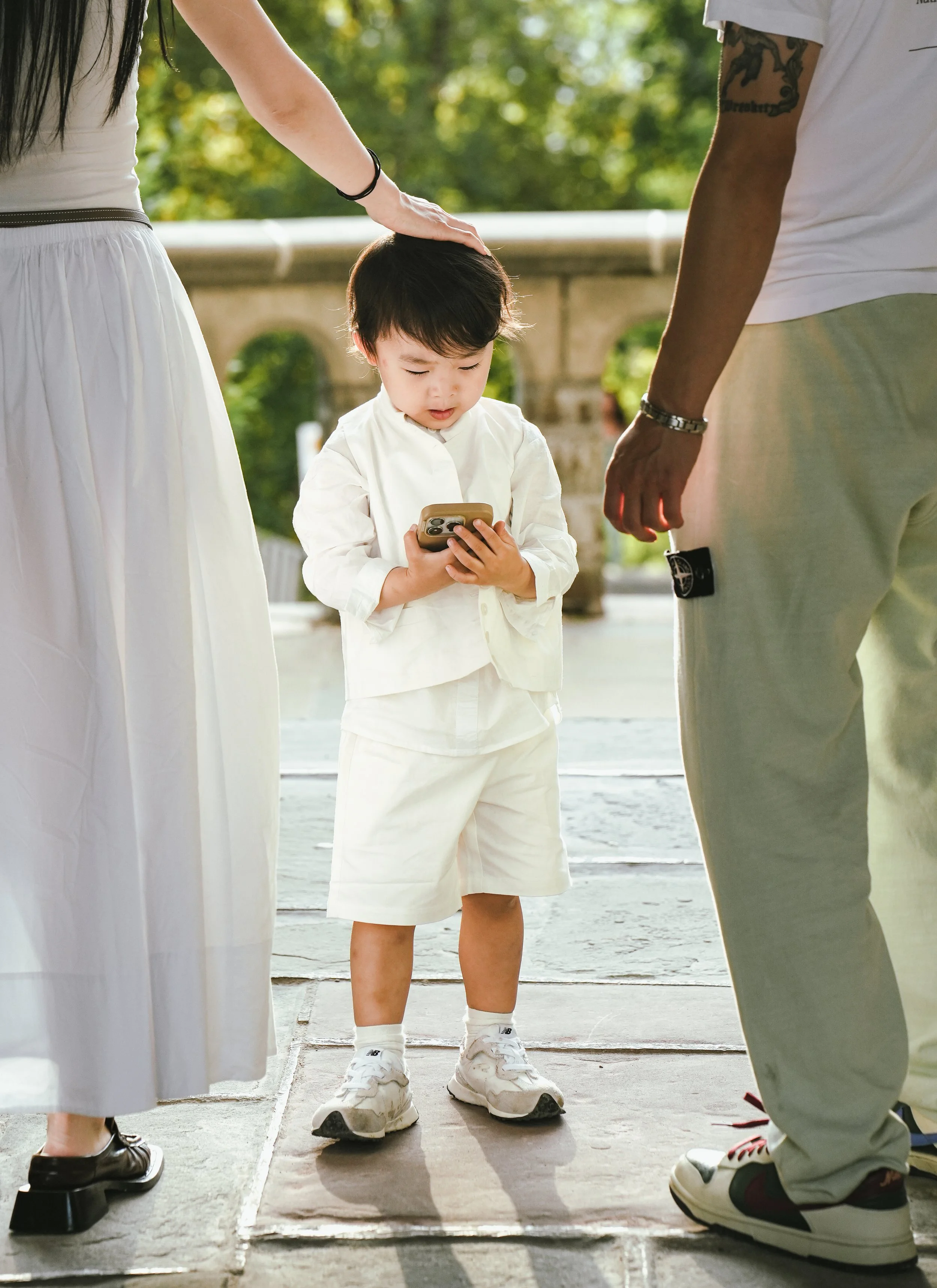 A young boy dressed in white standing on a skateboard while surrounded by three adults, one touching his head, outdoors with green trees and bright sunlight in the background.