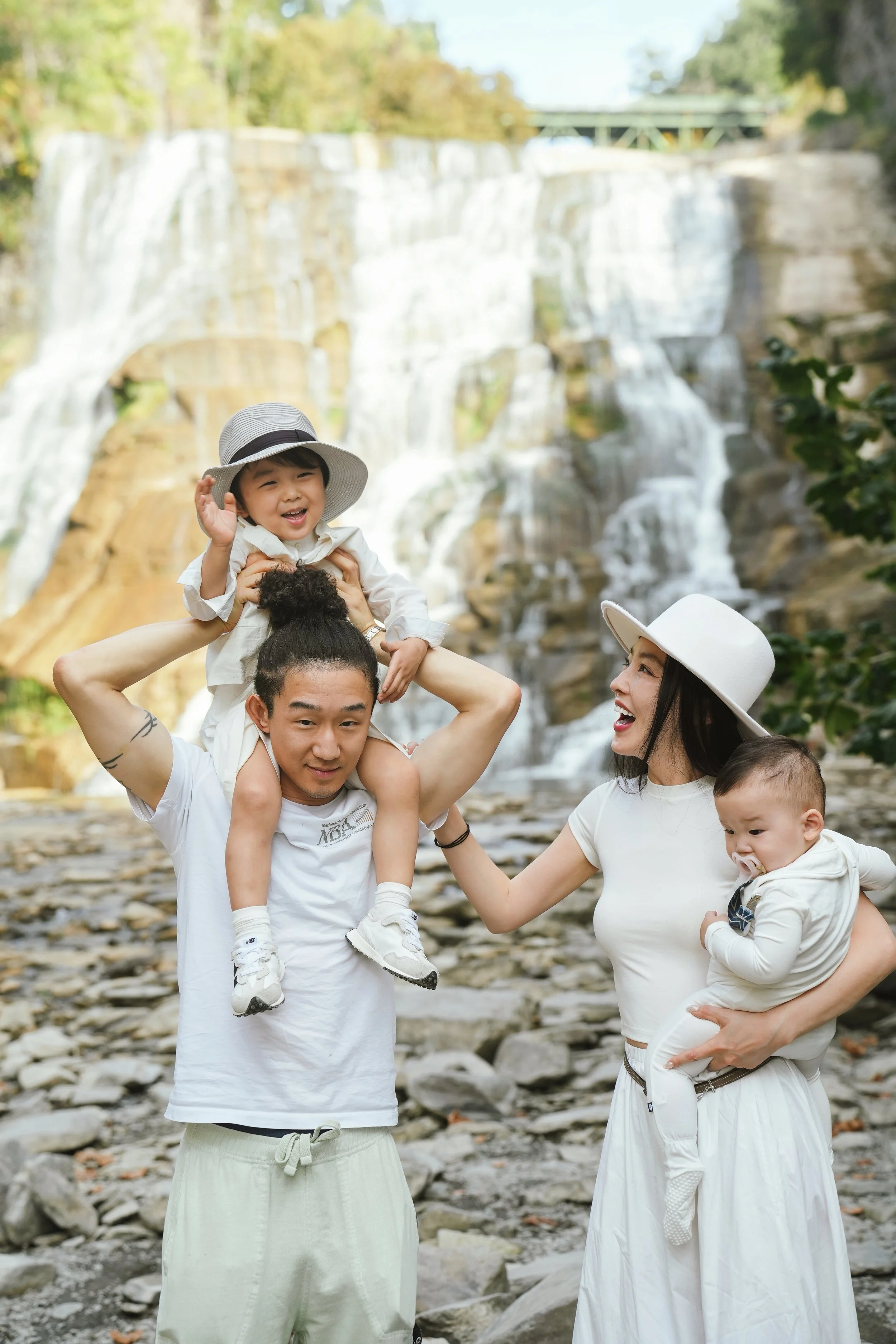 A family of four enjoying a day outdoors with waterfalls and trees in the background. The father is holding a young boy on his shoulders, and the mother is holding a young girl with a pacifier, while smiling and dressed in white.