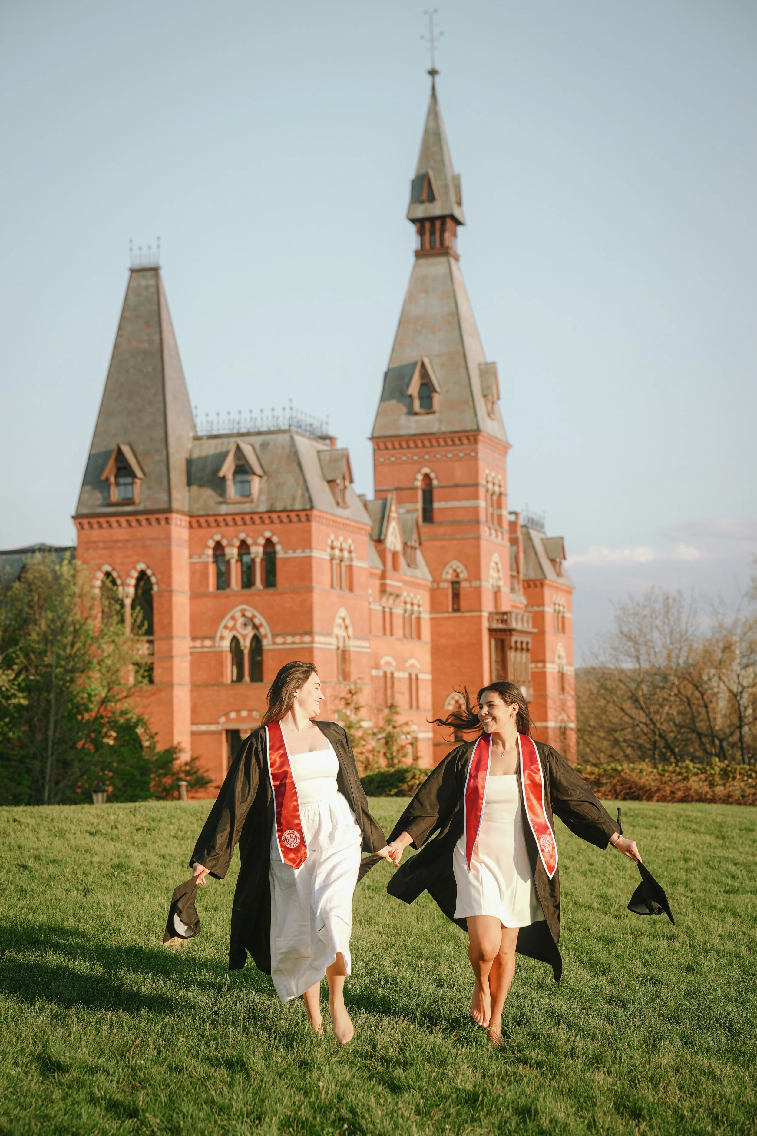 Two women in graduation gowns and stoles holding hands and walking barefoot on the grass, with a red-brick historic building with steeples in the background under a clear sky.