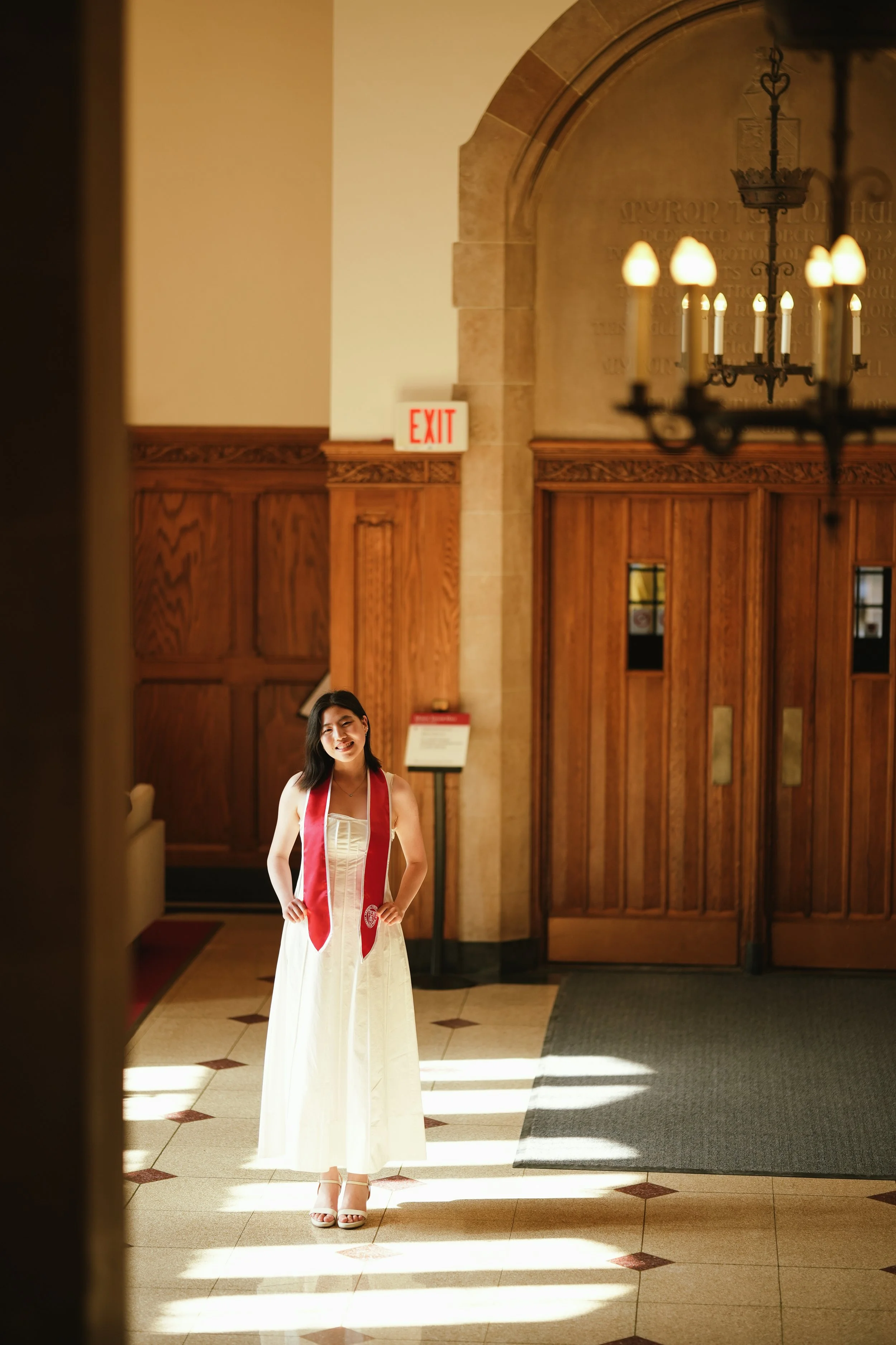 A young woman wearing a white dress, white high-heeled sandals, and a red stole stands in a church or cathedral lobby with wooden doors, ornate woodwork, and a chandelier. Natural light illuminates her, and an exit sign is visible on the wall behind 