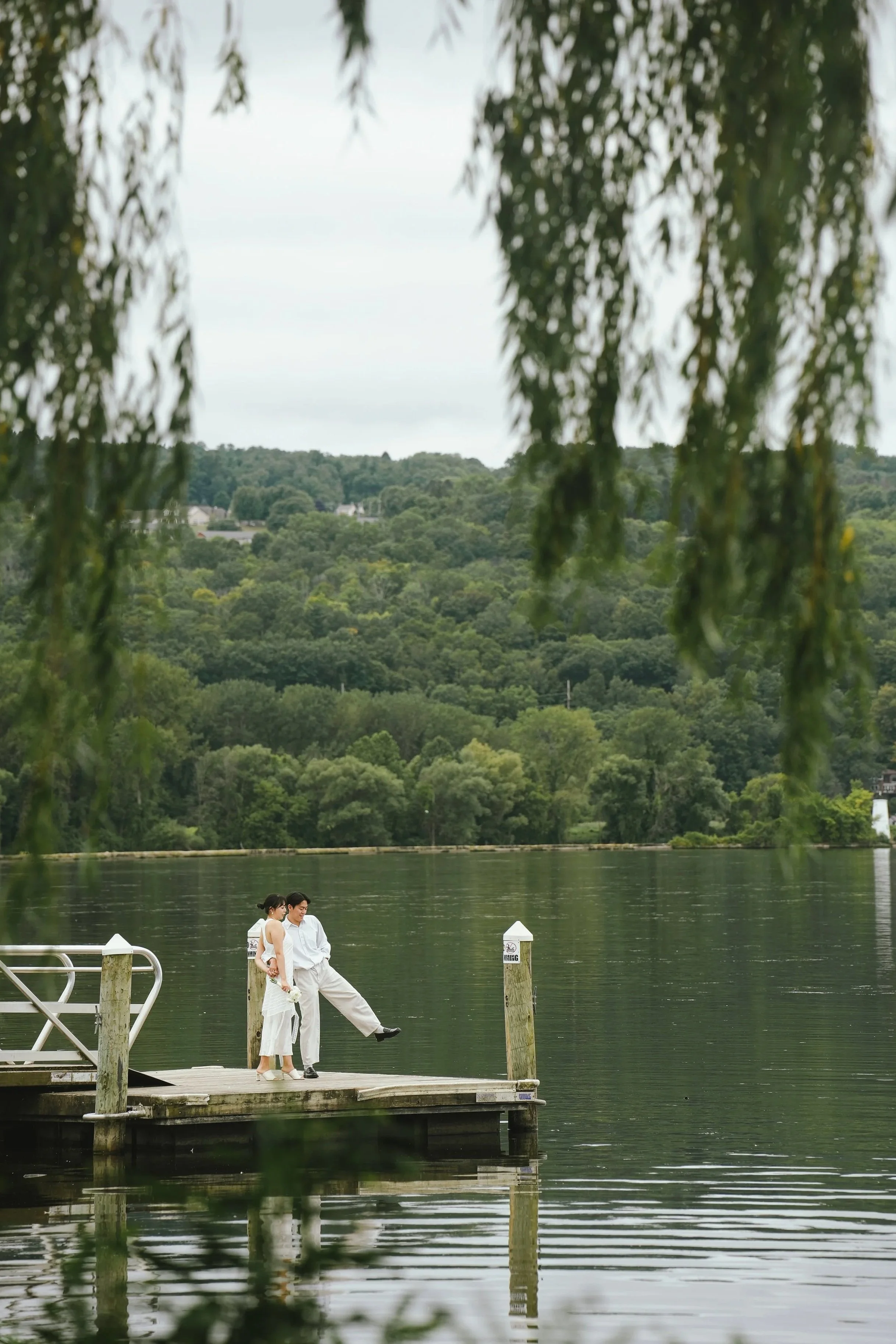 A couple dressed in white standing on a wooden pier by a lake, surrounded by trees and greenery, with a hilly landscape in the background, overcast sky, and hanging foliage in the foreground.