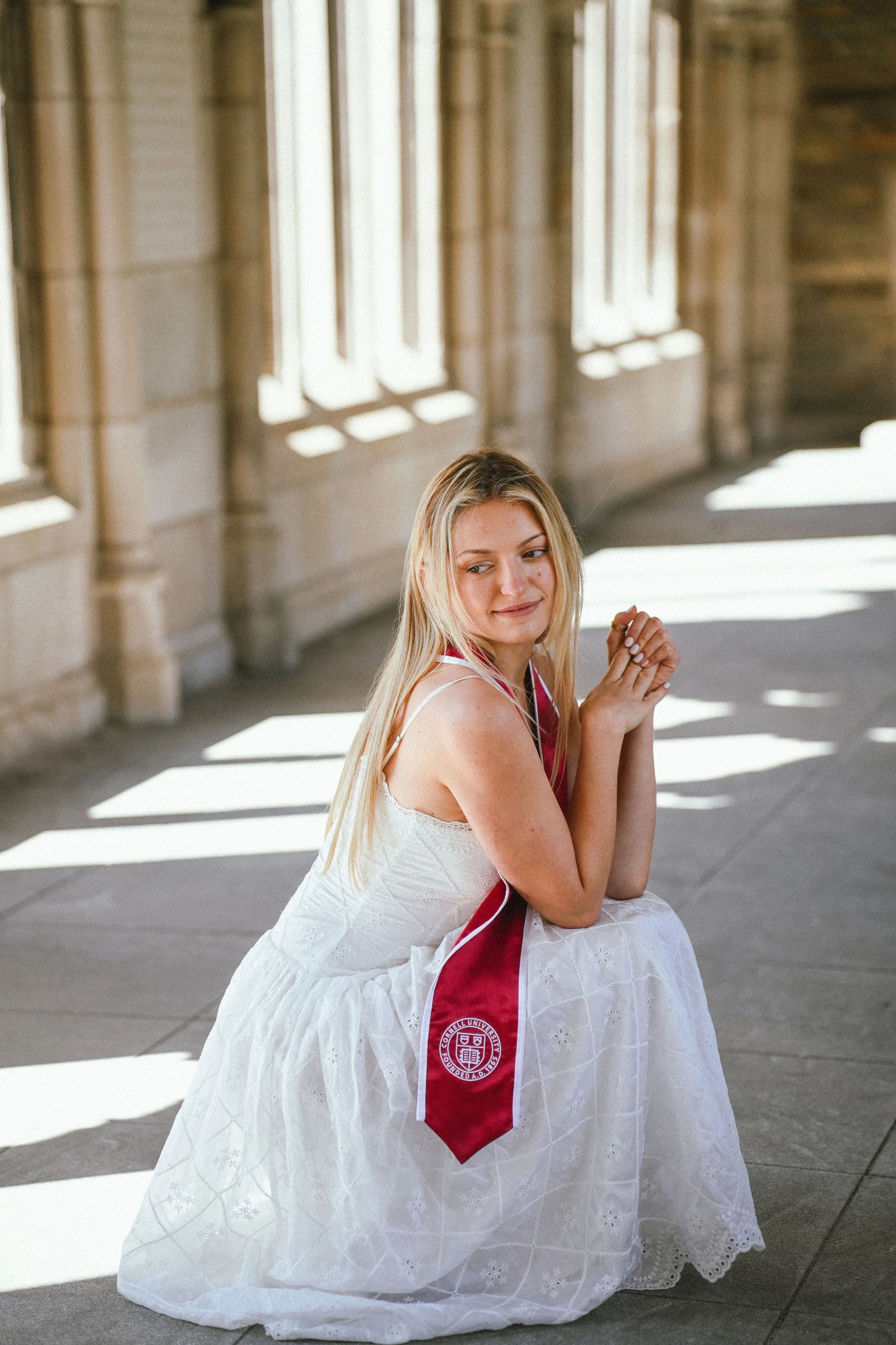 A young woman with long blonde hair sitting outdoors on a concrete surface, wearing a white dress with eyelet details and a red sash, with a Cornell University emblem on it. She is smiling softly, with her hands clasped together near her face, and su