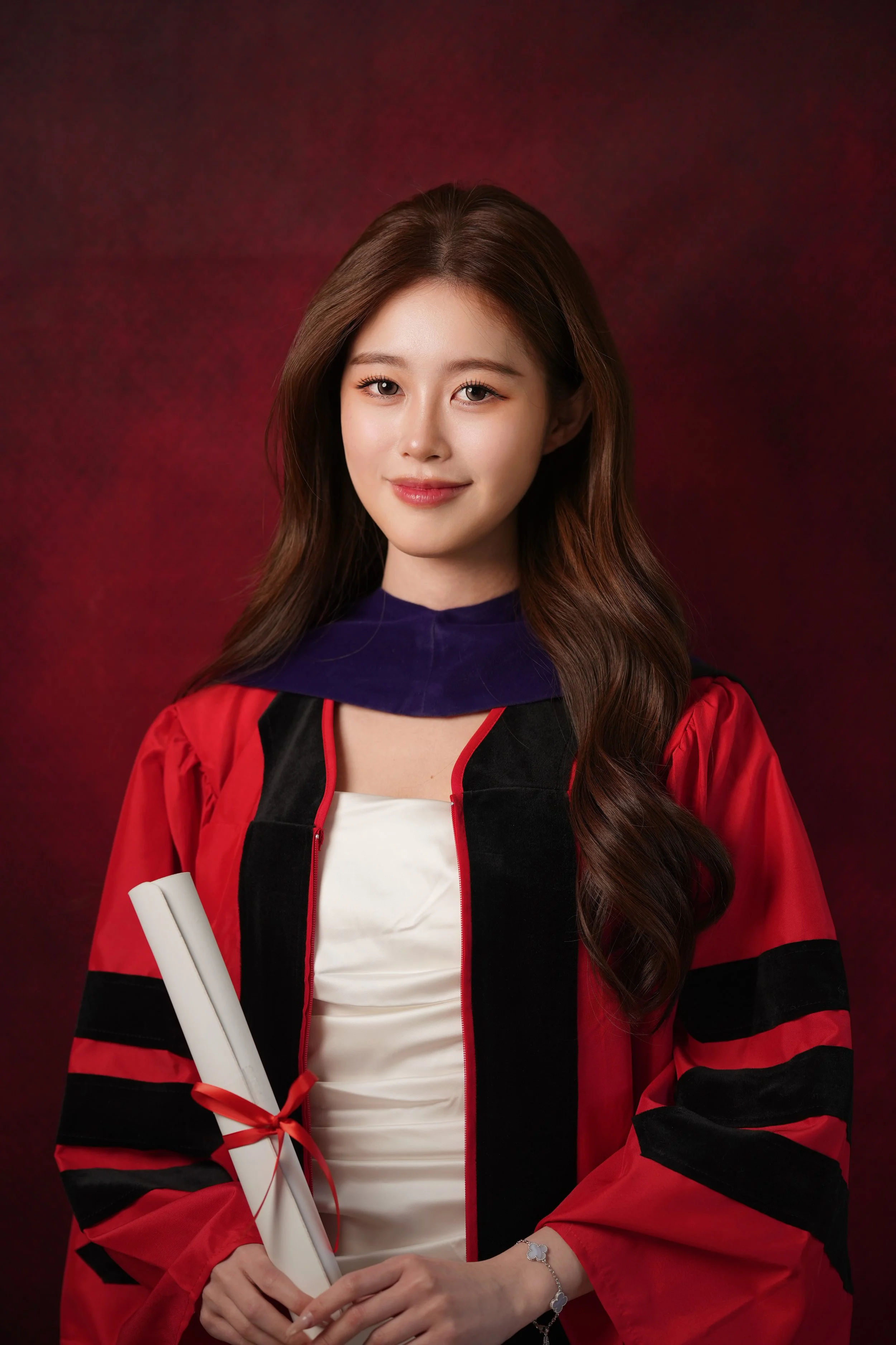 A young woman with long wavy brown hair, wearing a red and black graduation gown and holding a rolled white diploma tied with a red ribbon, standing against a dark red background.