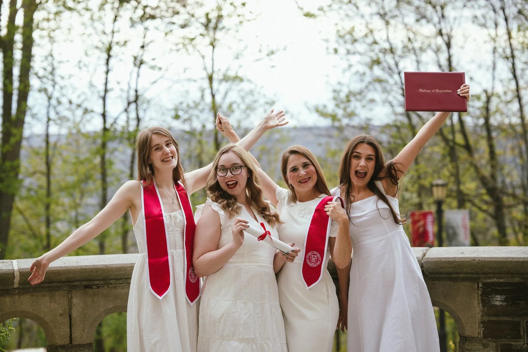 Four women celebrating graduation outdoors, wearing white dresses and red stoles, some holding diplomas, with trees in the background.