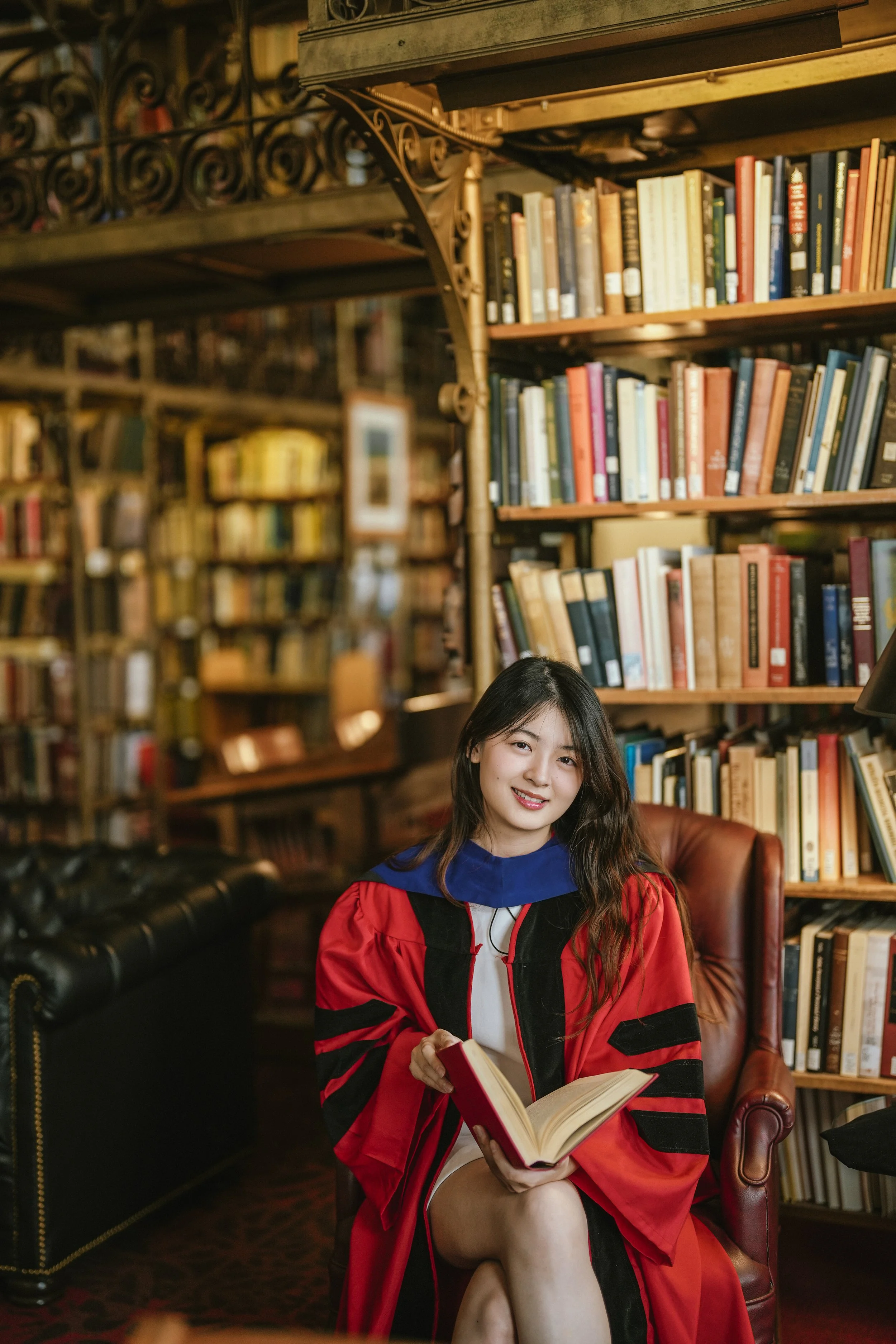 A young woman in a red and black graduation gown sitting on a leather armchair in a library, holding an open book and smiling.