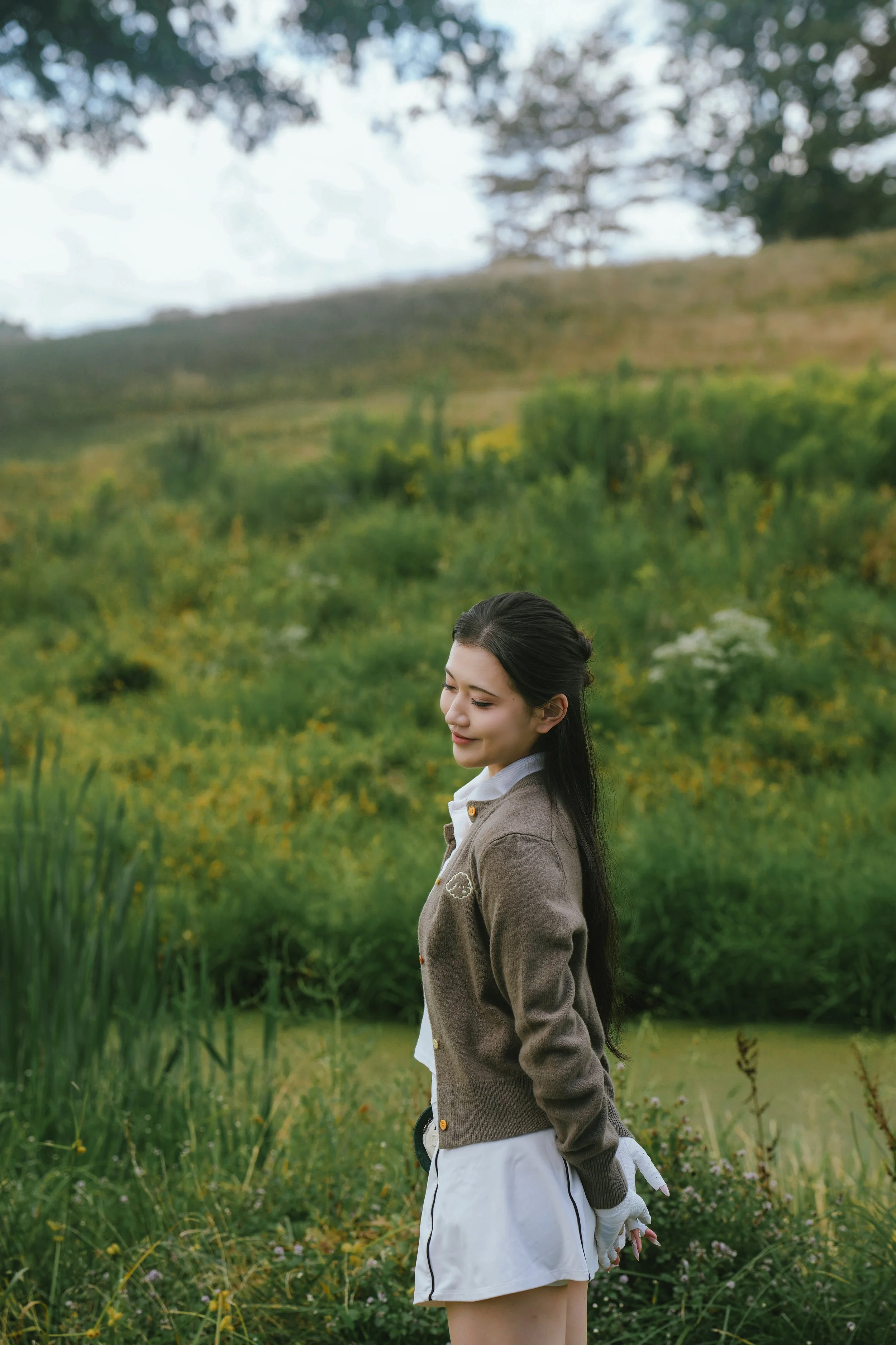 A woman with long dark hair standing in a lush green field near a pond with her eyes closed, wearing a gray cardigan and white skirt, with a peaceful expression.
