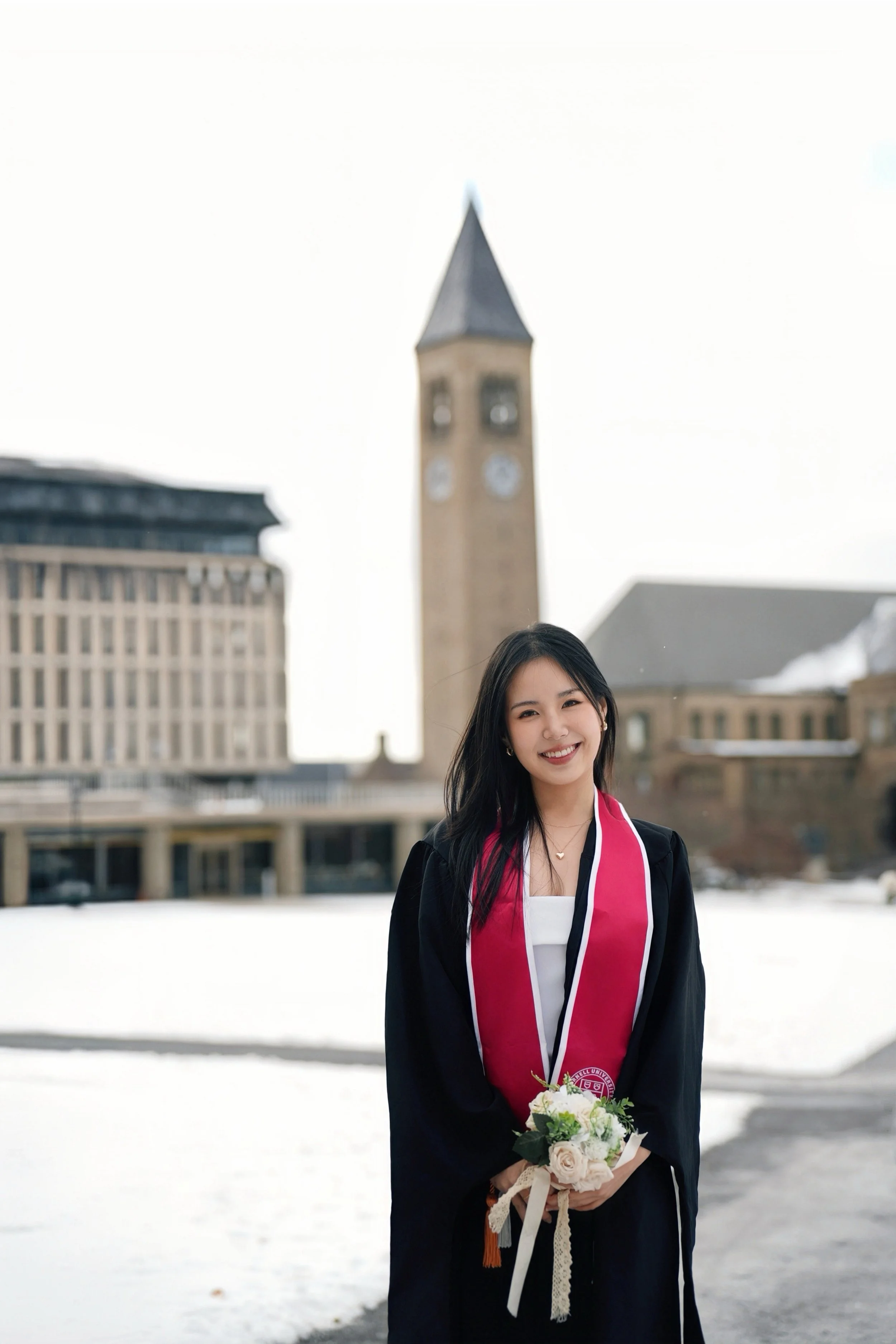 Young woman in graduation gown and pink stole holding a bouquet of flowers, standing outdoors with a clock tower and buildings in the background on a snowy day.