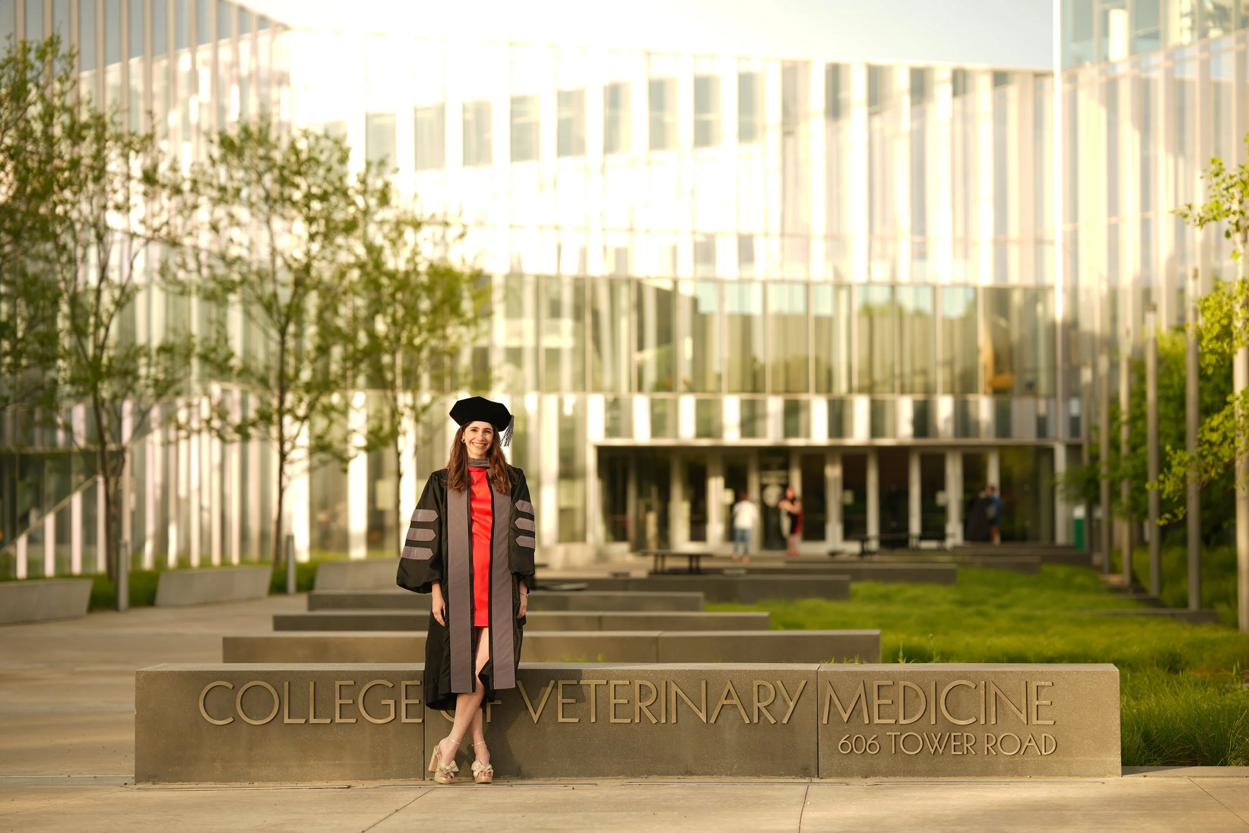 A woman in graduation attire, including a black gown and a cap, standing in front of a college building with the words 'College of Veterinary Medicine' engraved on a stone sign. The background features a modern glass building and greenery.