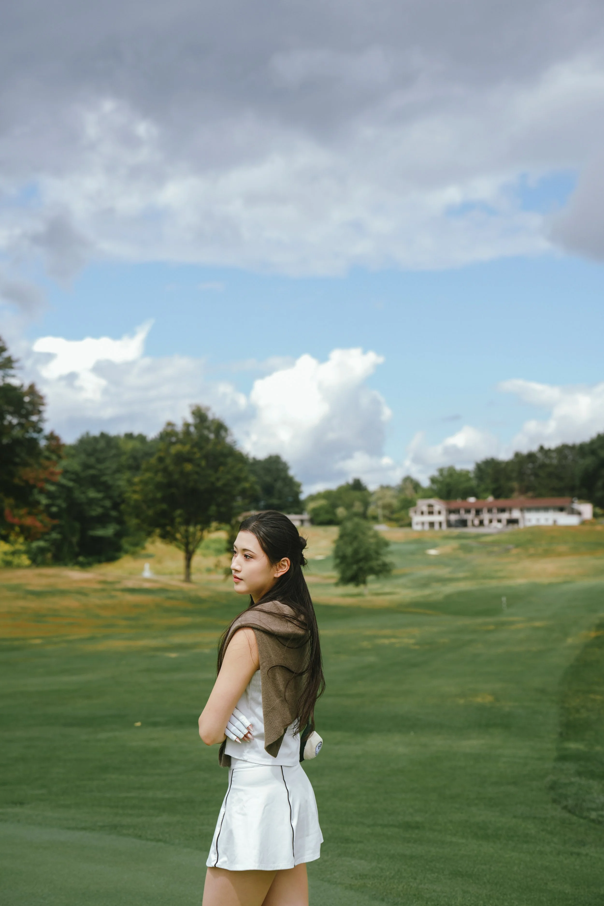 A young woman with long dark hair standing on a golf course with her arms crossed, looking to the side, with trees and a building in the background under a partly cloudy sky.