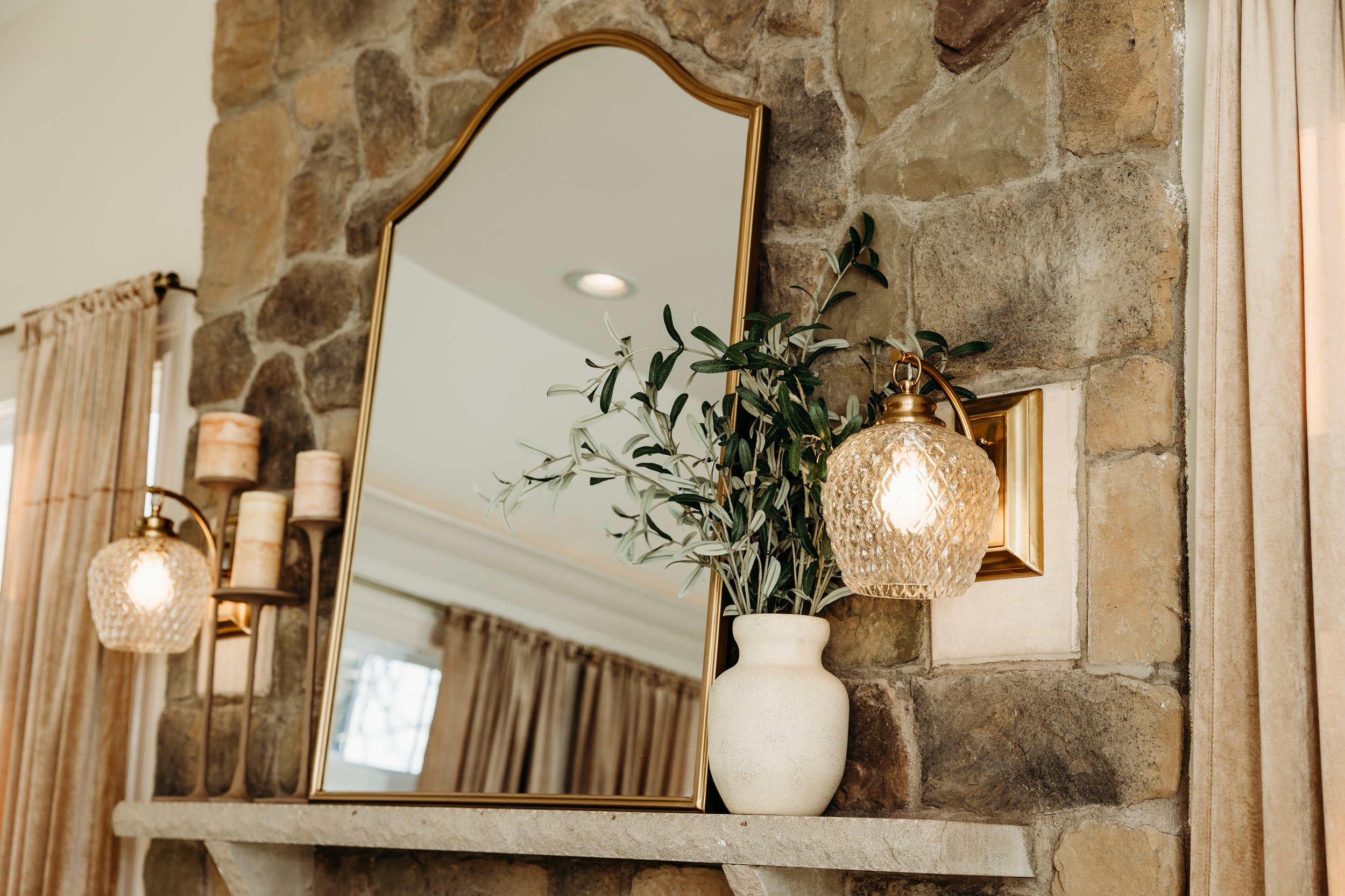 Cozy rustic decor with a wall mirror, a plant in a vase, candles, and a decorative sconce light on a stone wall.