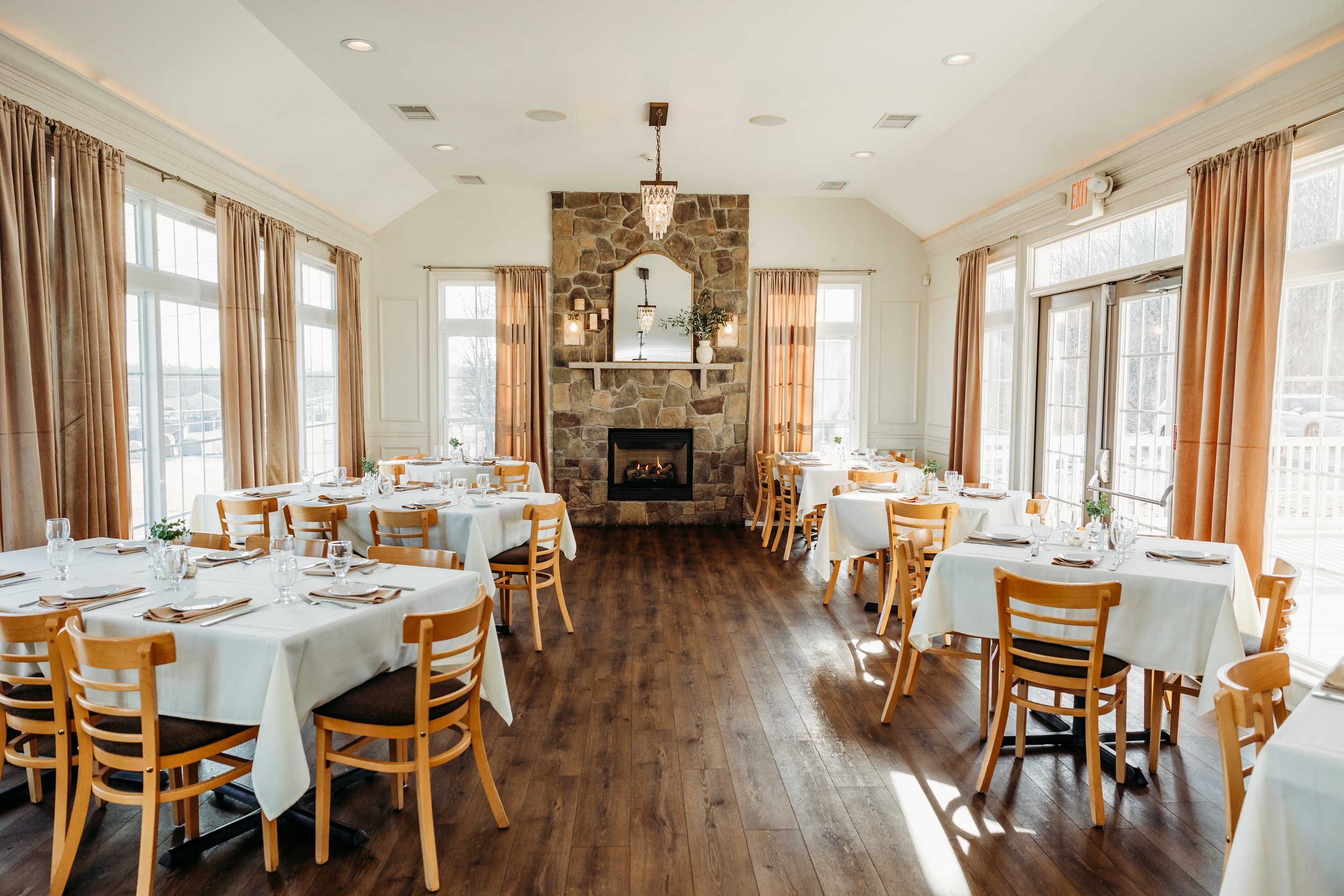 Elegant restaurant interior with wooden flooring, tables covered with white tablecloths, light wooden chairs, large windows with curtains, and a stone fireplace.