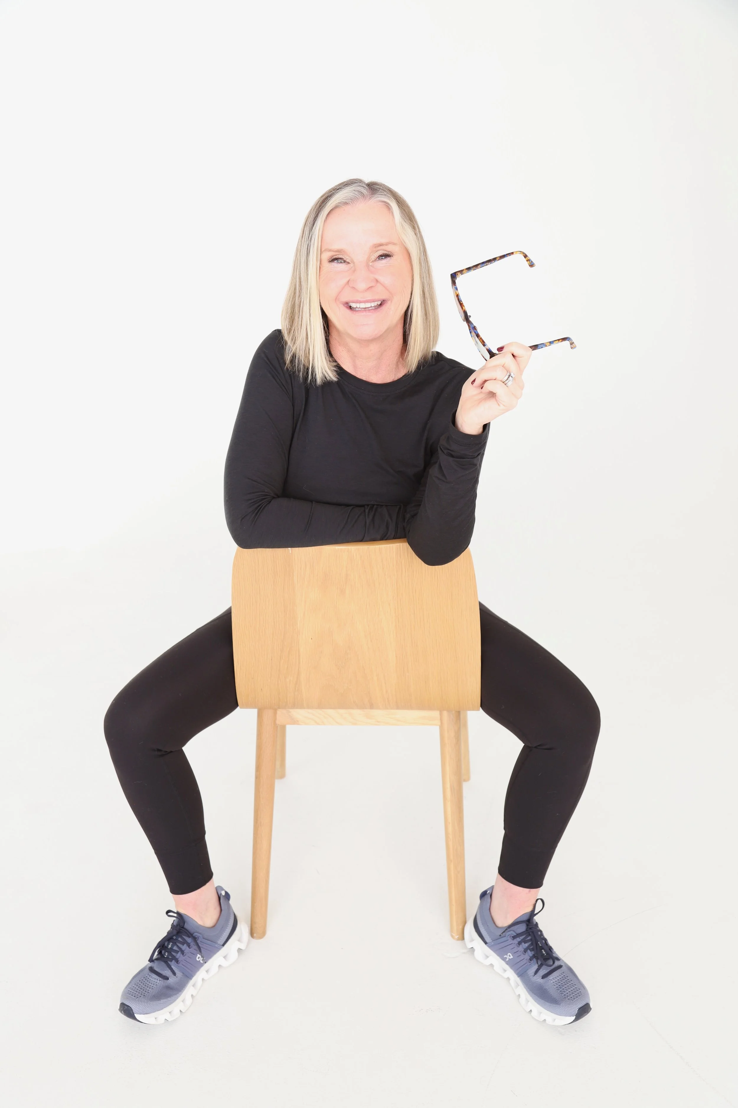 Woman sitting on a wooden chair with her legs spread, smiling, holding glasses in her right hand, wearing a black long sleeve shirt, black pants, and gray sneakers against a plain white background.