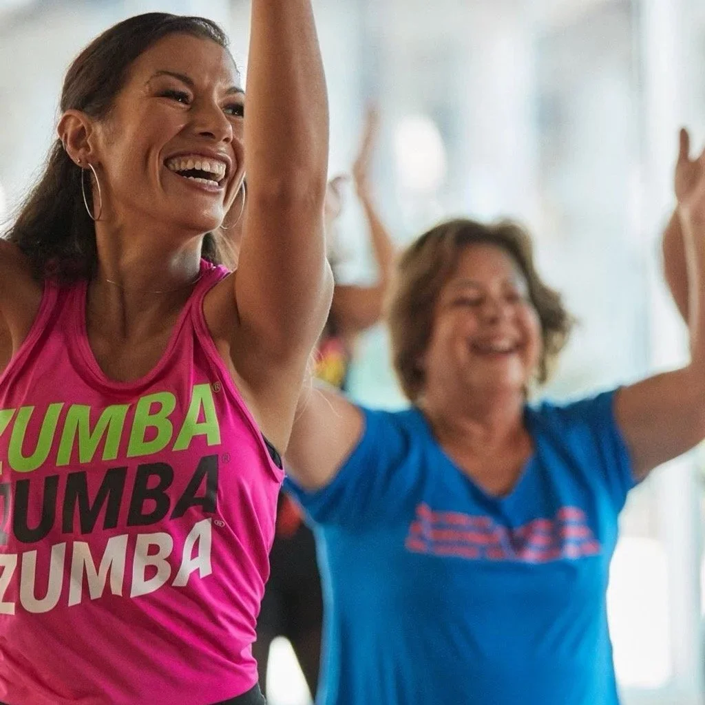 Eine fröhliche Frau in einem Zumba-Kurs mit ausgestreckten Armen, lachend, im Vordergrund. Im Hintergrund weitere Frauen, die ebenfalls tanzen, in einem Raum mit großen Fenstern, mit Zumba-Text oben rechts.
