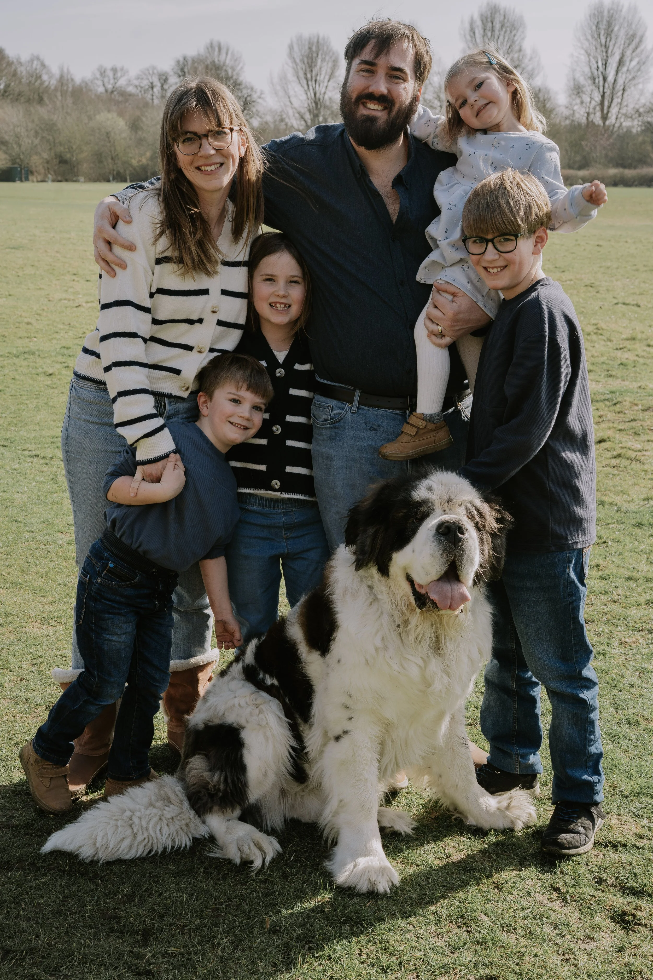 A happy family of six with a dog in a park. The family members are smiling, with the father holding a young girl. The dog, a large black and white Saint Bernard, sits on the grass in front.
