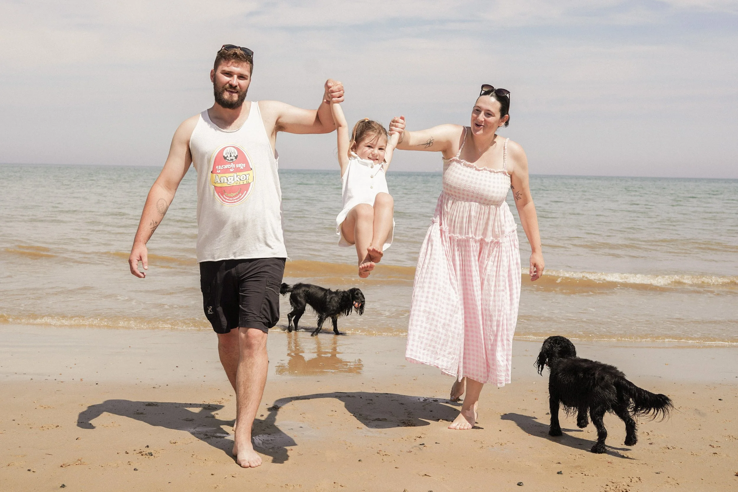 A family of four, with two dogs, enjoying a day at the beach. The father and mother are holding up a young girl between them, who is smiling. The father is wearing a tank top and shorts, and the mother is wearing a pink dress. The two dogs are black, one on the sand and the other near the water.