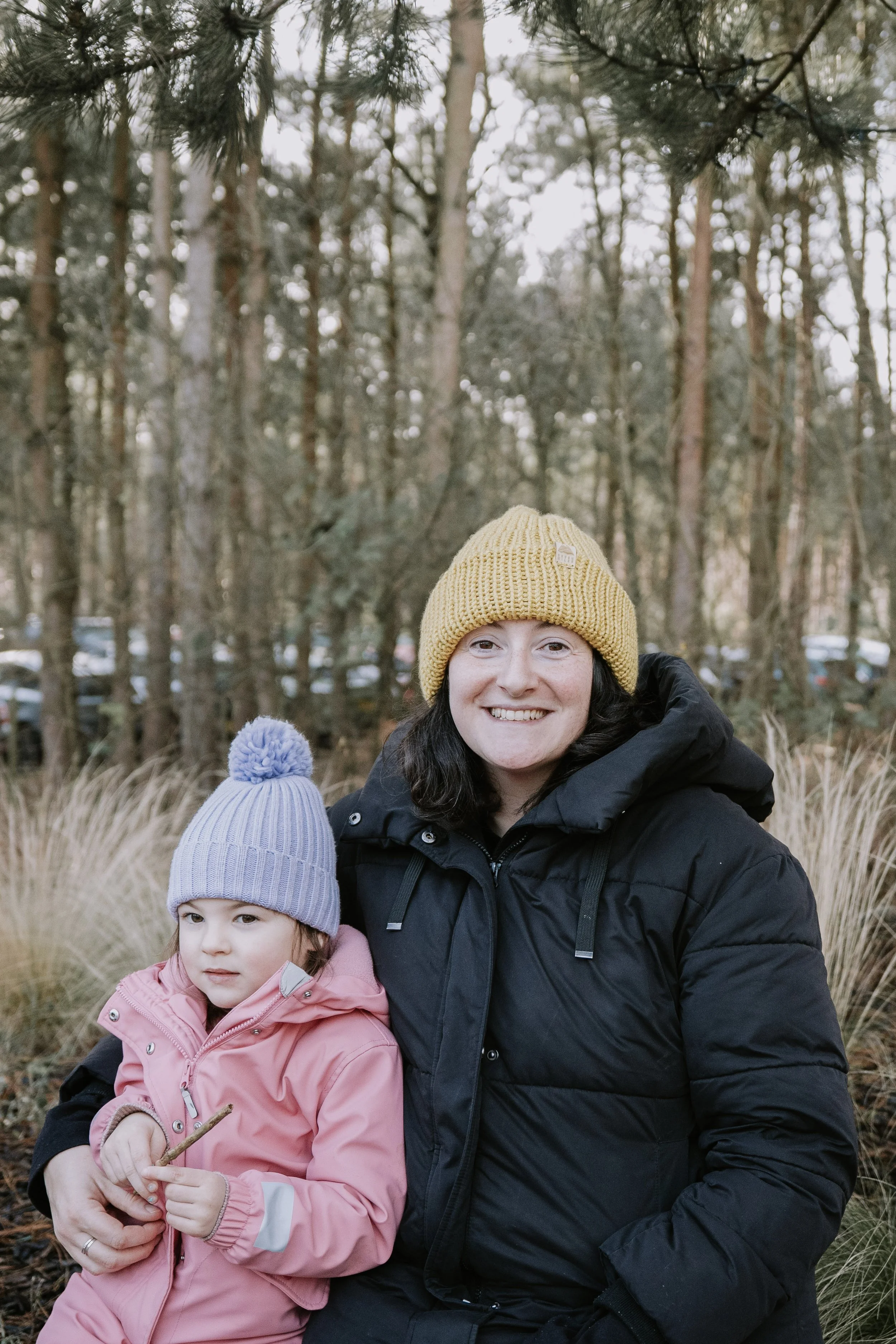 A woman and young girl outdoors in winter, both wearing knit hats and jackets, standing among trees and tall grass, smiling and posing for the camera.