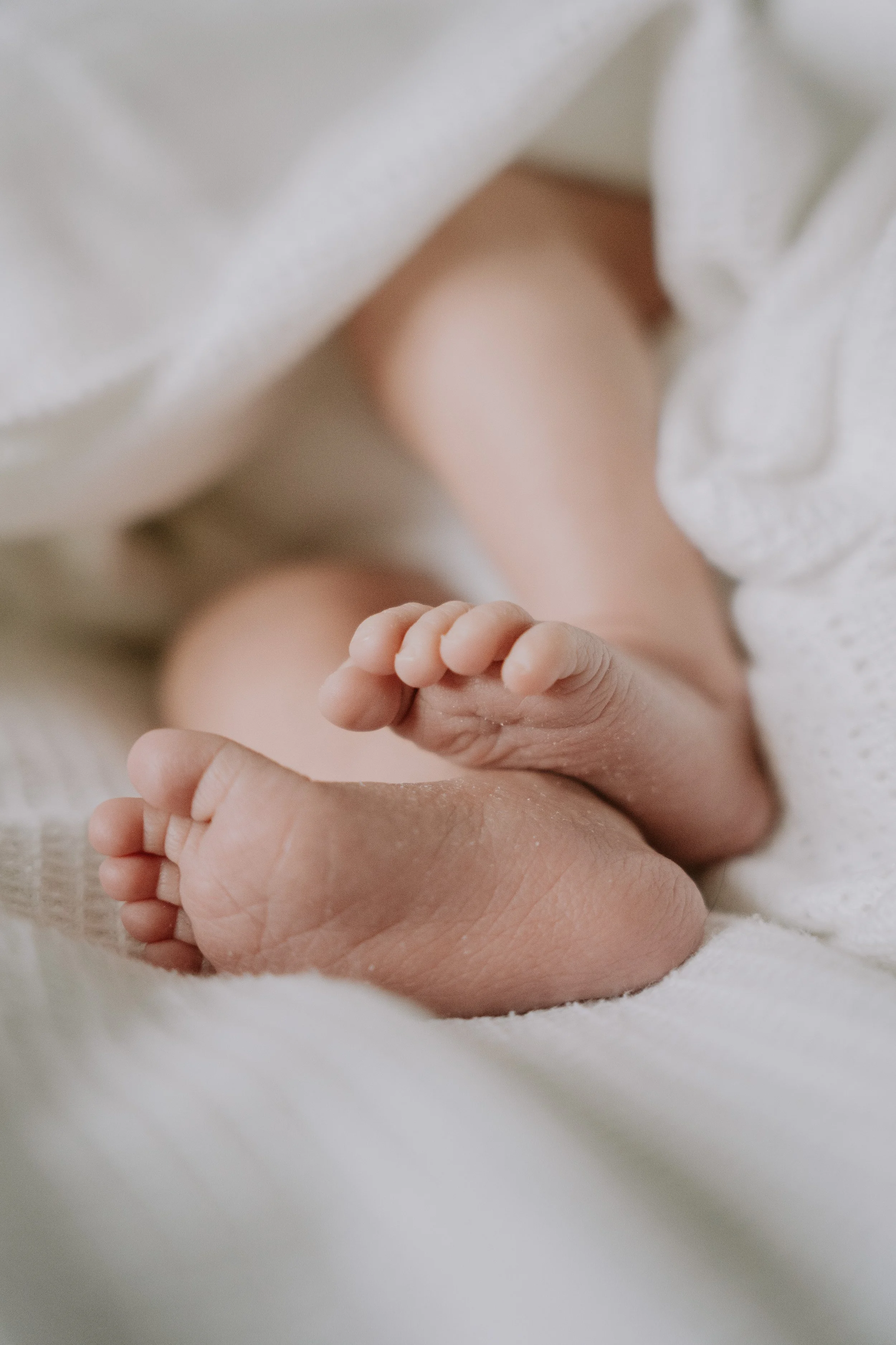 Close-up of a newborn baby's tiny hand grasping an adult's finger, both resting on a soft surface, with the baby wearing a white garment.