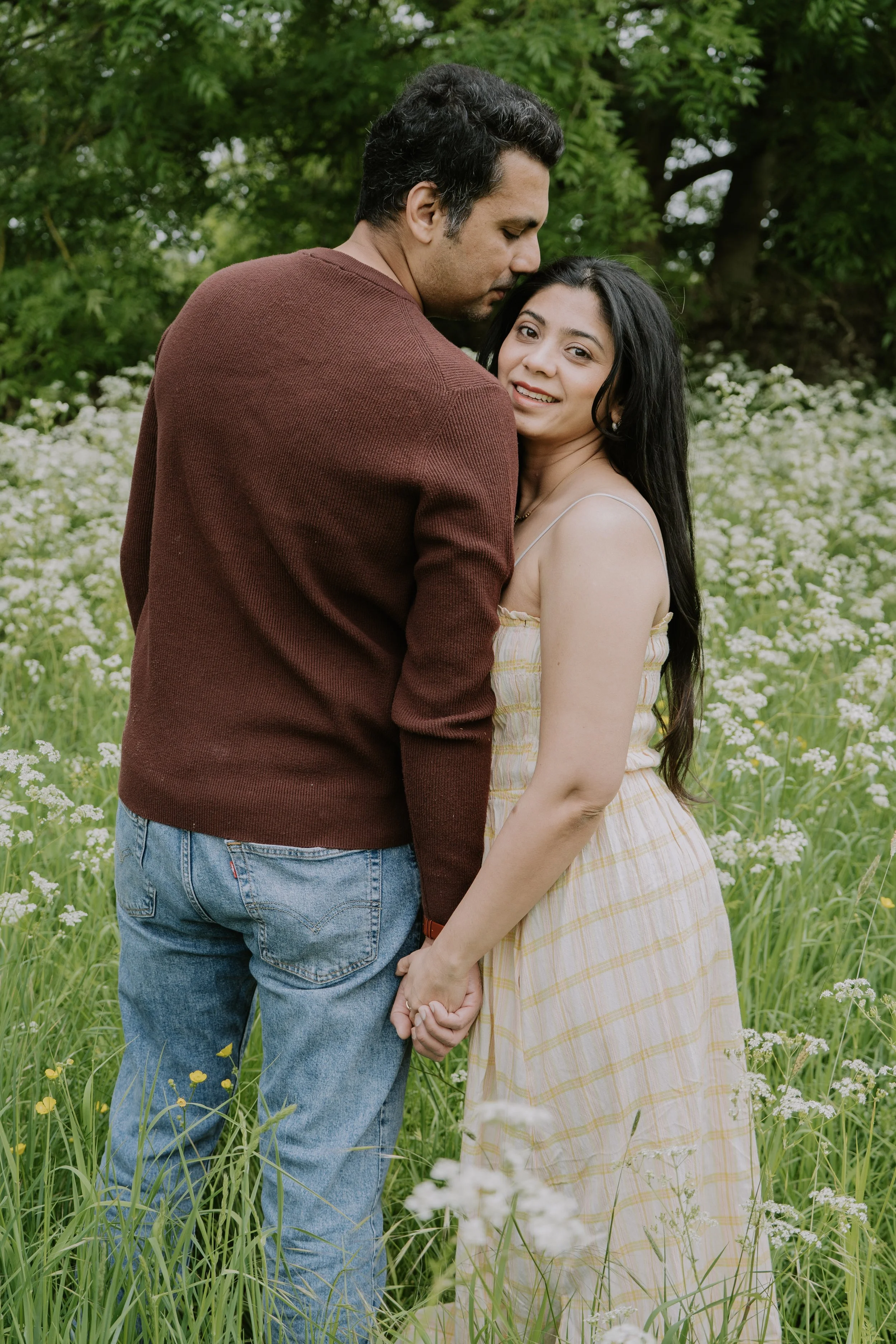 A couple holding hands in a field of white flowers, with green trees in the background, embracing each other lovingly.